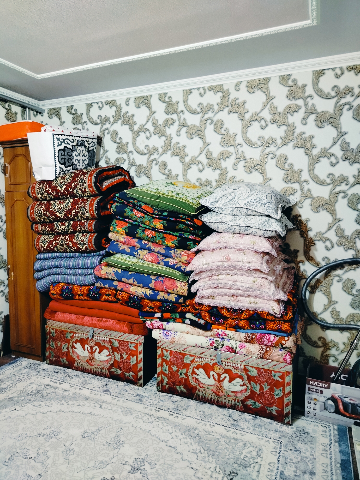 The image shows a room in a residential setting in the city of Kölkent, Kazakhstan. The room is decorated with a patterned wallpaper. A large stack of folded textiles, including blankets and pillows, is centrally placed. These textiles are colorful and feature various patterns, such as floral designs and stripes. Two decorative storage boxes with intricate red fabric covers, each depicting white swans and floral motifs, are situated at the bottom of the textile stack. To the left, a portion of a wooden wardrobe or cabinet is visible. A vacuum cleaner and its box are present to the right. The floor is covered with a light-colored rug featuring a subtle pattern. The scene appears to be a domestic storage area within a home.