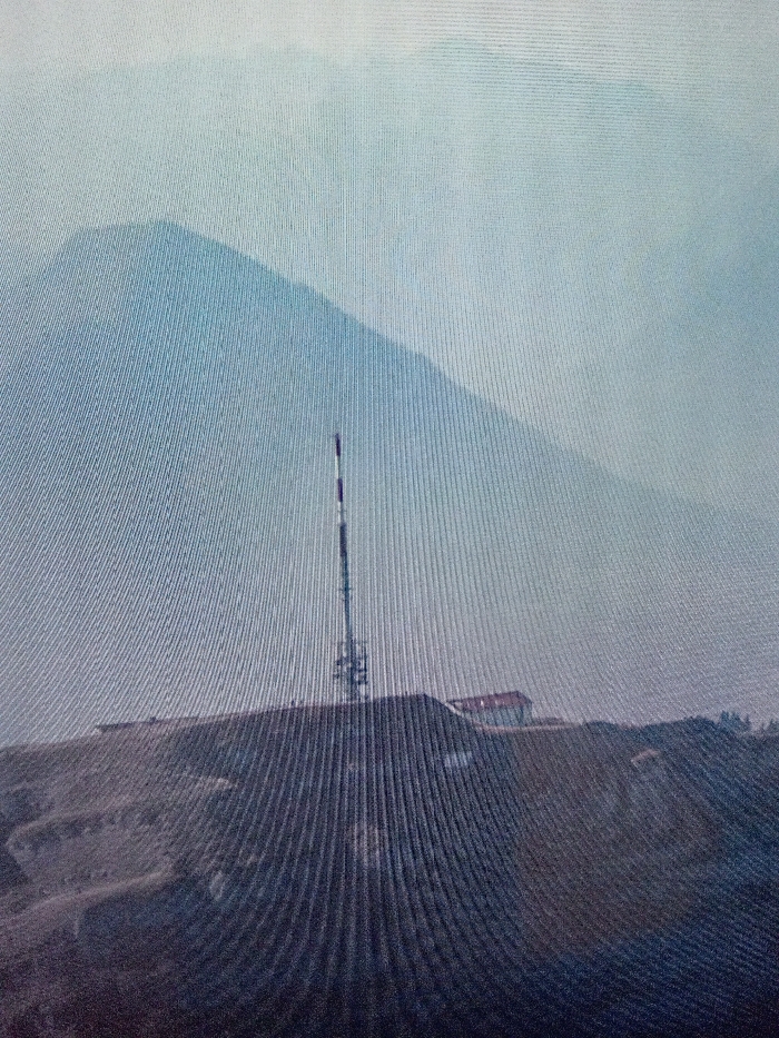 The image presents a daytime scene located in Ijoko, Nigeria, captured with visible vertical scan lines suggesting a photograph of a digital display. A tall communications tower, featuring a lattice construction with an upper section painted in alternating white and dark bands, stands centrally on an elevated, arid-appearing landmass. Behind the tower, to its right, a single-story structure with a distinct reddish roof is visible on the same elevation. The background features a large, indistinct mountain or elevated terrain, obscured by atmospheric haze against a light, diffuse sky. No individuals or specific actions are discernible within the frame.