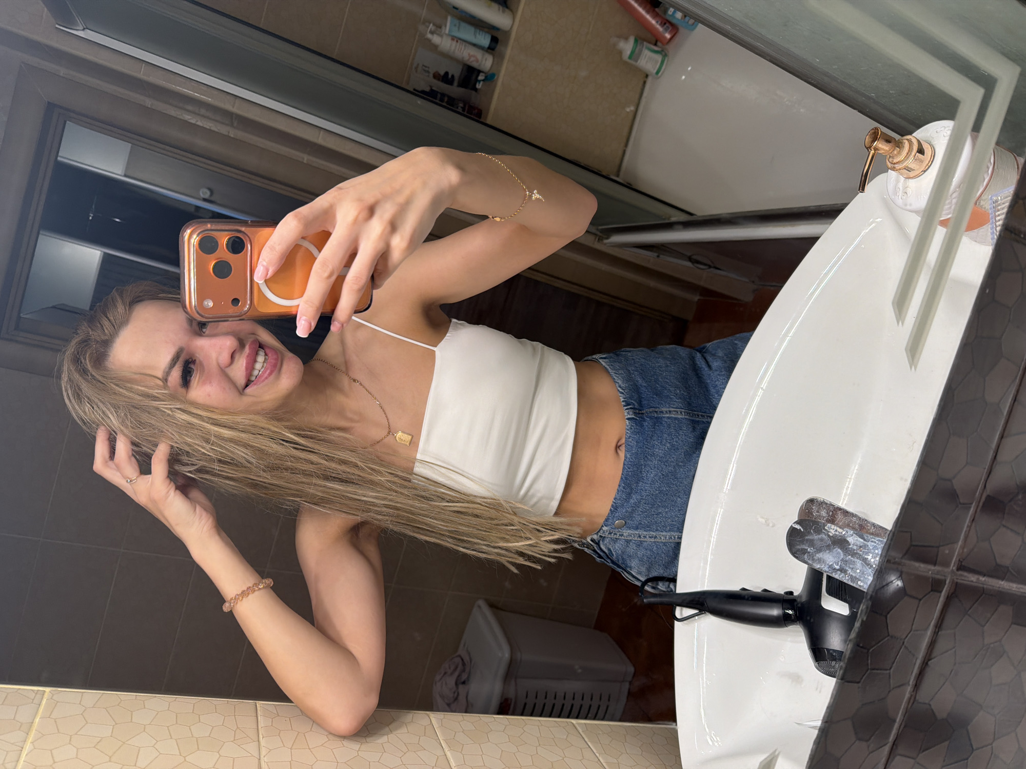 A young woman with blonde hair is taking a selfie in a bathroom, holding an orange-cased smartphone in her right hand and touching her hair with her left. She is wearing a white crop top and blue denim shorts. The bathroom has tiled walls and a sink area with a black hairdryer and a soap dispenser visible. The scene appears to be in a residential bathroom in Kyiv, Ukraine.