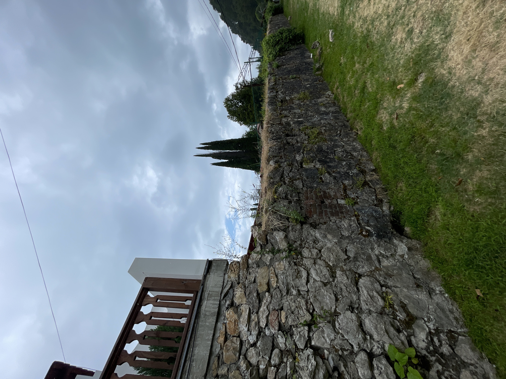 The image depicts an outdoor scene with a stone wall forming a prominent boundary. The wall is constructed from irregular, weathered stones of varying shades of grey and brown, with some patches of green moss and vegetation growing on its surface. To the right of the wall, a well-maintained grassy area slopes upwards, with some dry, yellowed patches mixed in with the vibrant green.
Beyond the stone wall, several tall, slender cypress trees stand out against a cloudy sky. In the background, a tree-covered hillside is visible, suggesting a mountainous or hilly terrain. Power lines are also present, stretching across the sky.
In the foreground, on the left side of the image, a section of a wooden railing and a white-capped structure are visible, indicating the presence of a building or balcony. The wooden railing has an intricate, decorative pattern.
The weather appears overcast, with a thick layer of grey clouds covering the sky, suggesting it might be a cloudy day or approaching rain. The time of day is not clearly discernible due to the diffused light. The location context provided is Abaata, Abkhazia. There is no text visible in the image. The overall mood is one of a somewhat natural, possibly rural or semi-rural setting with hints of human habitation.