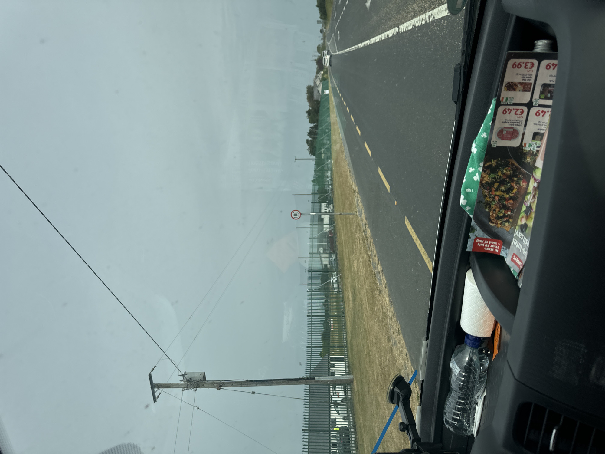 The image shows a view from inside a vehicle looking out at a road in Ireland. The sky is overcast and gray. The road is asphalt with yellow dashed lines marking the lanes. A speed limit sign with "100" in a red circle is visible on the right side of the road. On the left side of the road, there is a metal fence with a green mesh covering extending behind it. Beyond the fence, there are buildings and parked cars. The foreground of the image shows the interior of the vehicle, with a water bottle and a roll of white material placed in a compartment on the dashboard. There are also some promotional flyers visible in the compartment. A suction cup mount for a device is also visible in the lower left corner.
