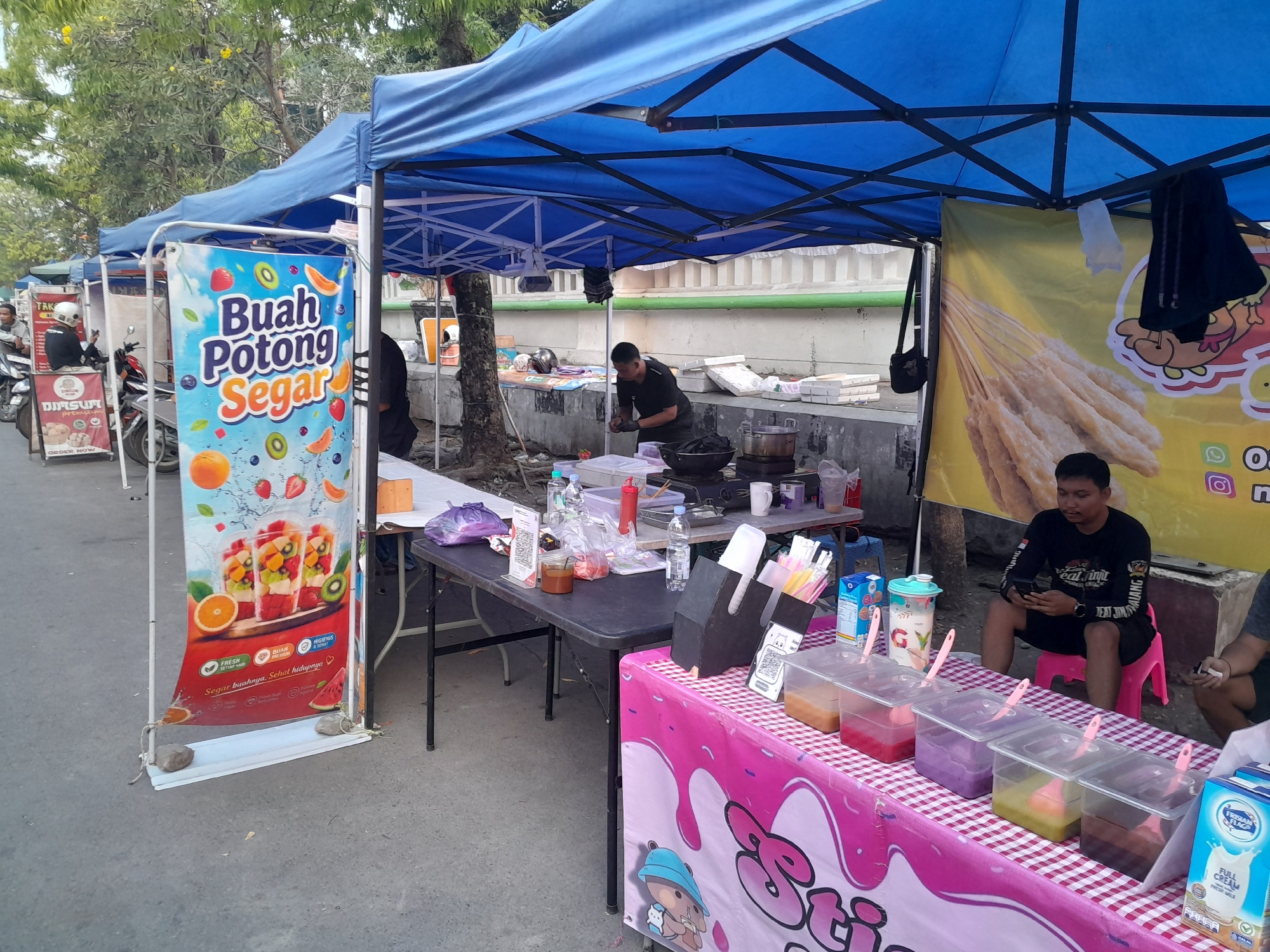 The image depicts an outdoor food stall, likely in Slawi, Indonesia, as suggested by visual cues and common street food setups. The stall is covered by a blue canopy tent. In the foreground, a table is laid out with a pink and white checkered tablecloth. On this table, several clear plastic containers hold brightly colored liquids, suggesting a variety of beverages or toppings. A blue carton of "Frisian Flag" milk is visible on the right side of the table. A man in a black long-sleeved shirt is seated on a pink stool, looking at his phone.
To the left of this main stall, another tented area is visible. A vertical banner advertising "Buah Potong Segar" (Fresh Cut Fruit) with images of fruit and fruit drinks is prominently displayed. A table in this section has various items, including bottles of water, containers of sauce, and stacks of white styrofoam boxes. Two individuals are partially visible in the background on motorcycles.
Further back, another person is seen preparing food near a cooking station with a wok. The environment suggests a busy street or market setting, with concrete pavement and trees visible. The overall scene captures the daily operations of a local food vendor in Indonesia.