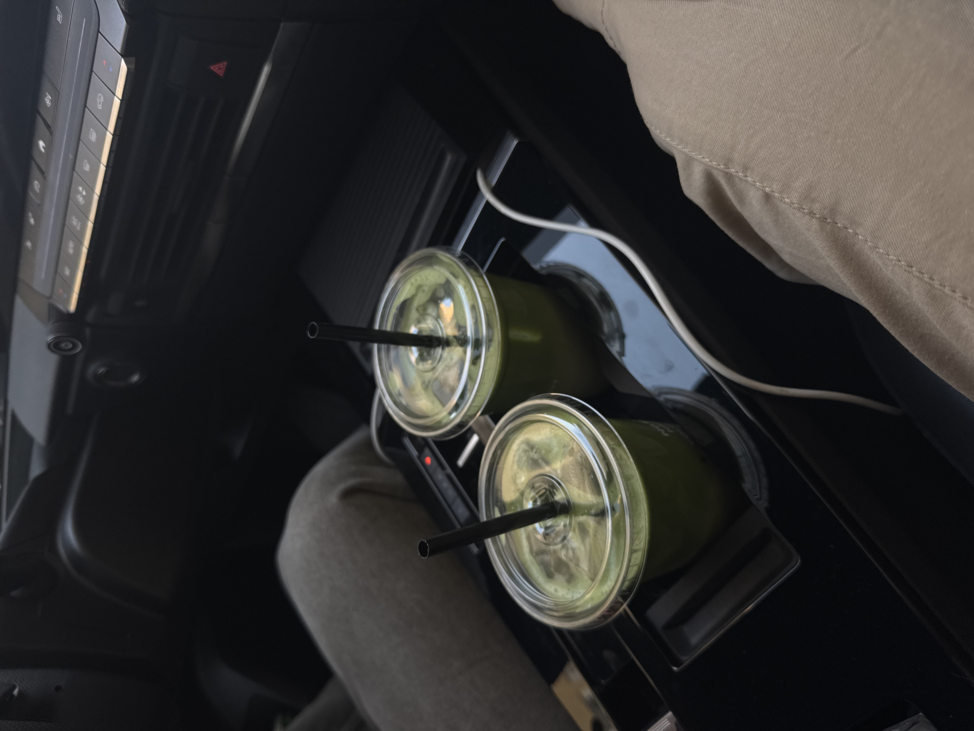 The image shows a car's interior with two cups of matcha latte in the cup holders. Each cup has a black straw. A white cable lies adjacent to the cups. The person on the right is wearing tan pants, and the person on the left is wearing grey sweatpants. The image was taken inside a car, potentially in transit, but it does not provide explicit information about the city. Therefore, there is no evidence for the location in Warsaw, Poland.