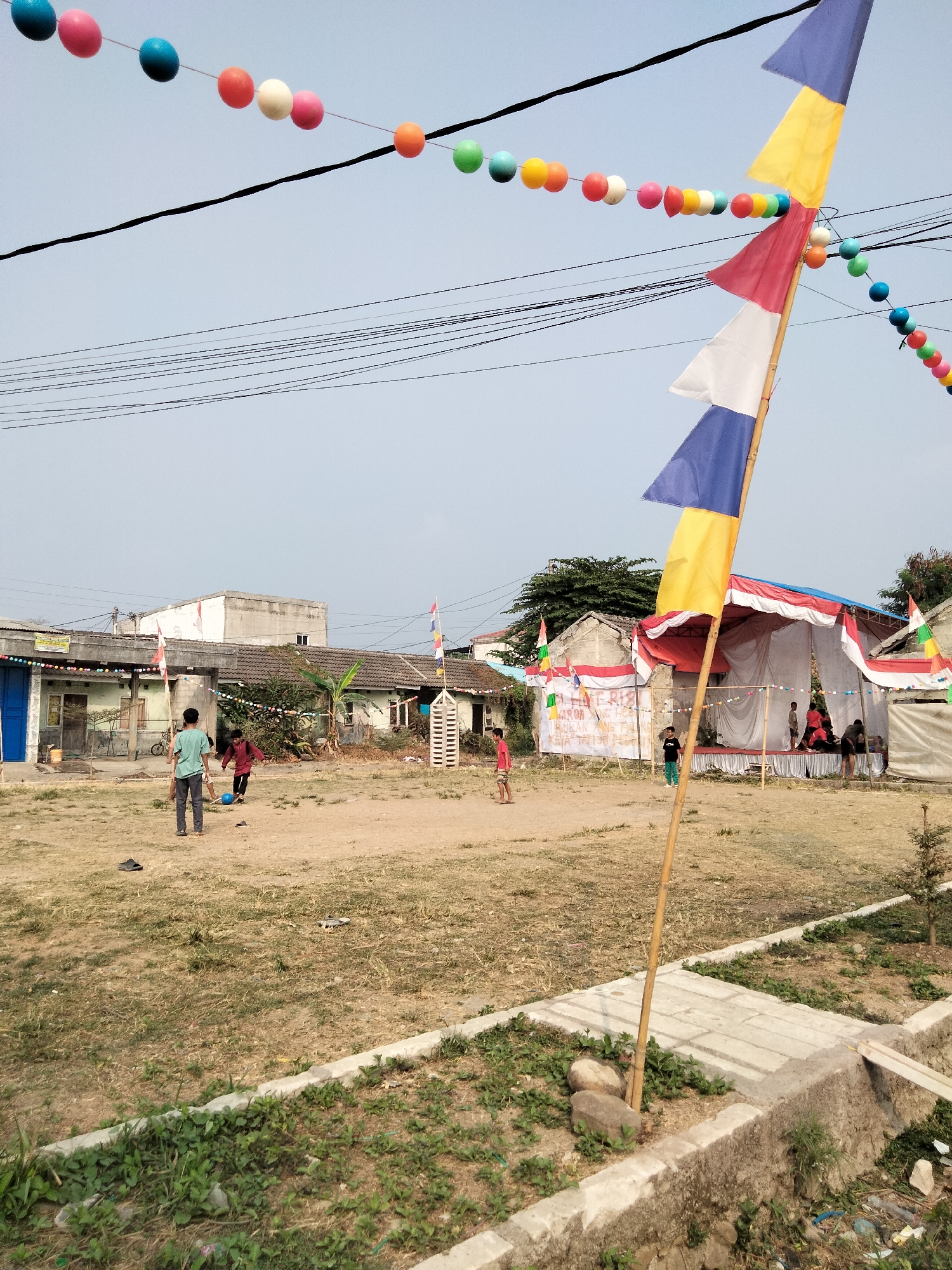 An outdoor scene is depicted in the city of Cariu, Indonesia, with a focus on a grassy field and the surrounding structures. In the foreground, a concrete barrier with some greenery and rocks is visible, with a bamboo pole planted next to it. The field itself is mostly dry grass with some patches of dirt, and a few scattered stones and debris.
In the mid-ground, several individuals are present. Two young males are engaged in a game of soccer, with one kicking a blue ball while the other watches. Another young male stands with his back to the camera, observing the game. In the background, a white tent with red and white drapes is set up, possibly for an event. Several people are gathered under and around the tent. Adjacent to the tent are buildings with tiled roofs, typical of residential structures in the area, and one building has a blue door. Overhead, electrical wires are strung across the sky, and strings of colorful round decorations, likely balloons or lanterns, are suspended between poles and wires. Indonesian flags, red and white, are also displayed on poles. The sky is clear and overcast.