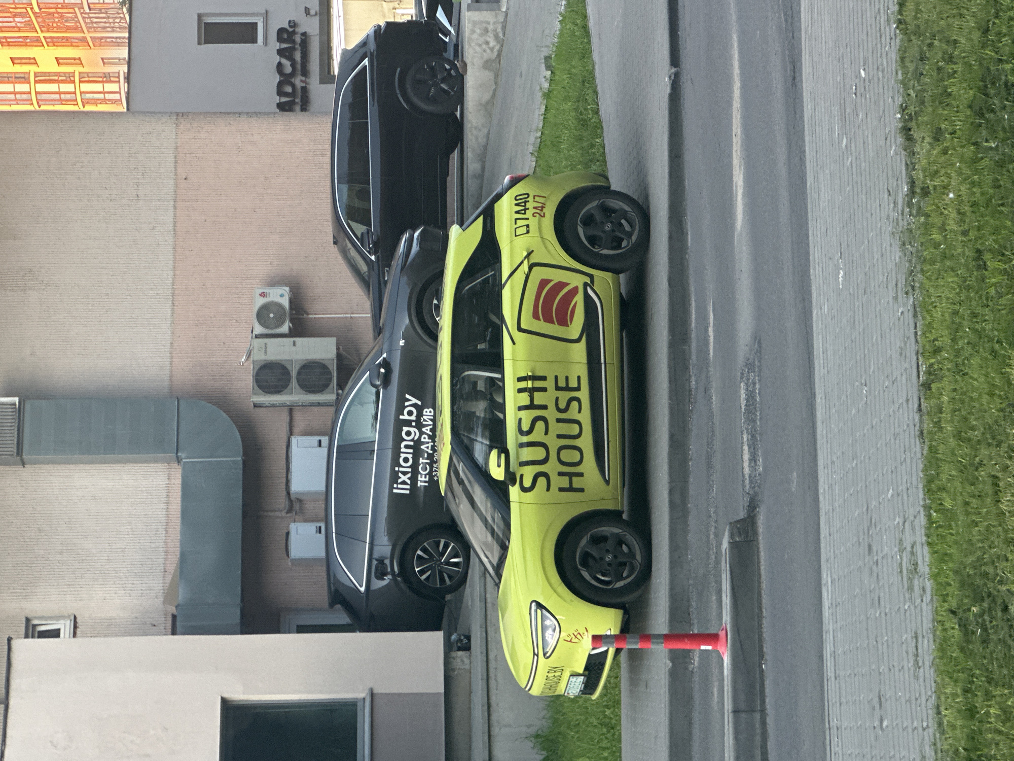 In an urban environment, a bright yellow electric car with "SUSHI HOUSE" written in large black letters on its side is parked on a sidewalk. The car features a stylized logo of sushi rolls and the phone number "7440 24/7" also prominently displayed. Behind the yellow car, two dark-colored SUVs are parked. The rear SUV has "lixiang.by" and "ТЕСТ-ДРАЙВ" written on its side. The buildings in the background are modern, with visible air conditioning units and architectural details. A red and white traffic cone stands in front of the yellow car. The scene is captured during daylight.
