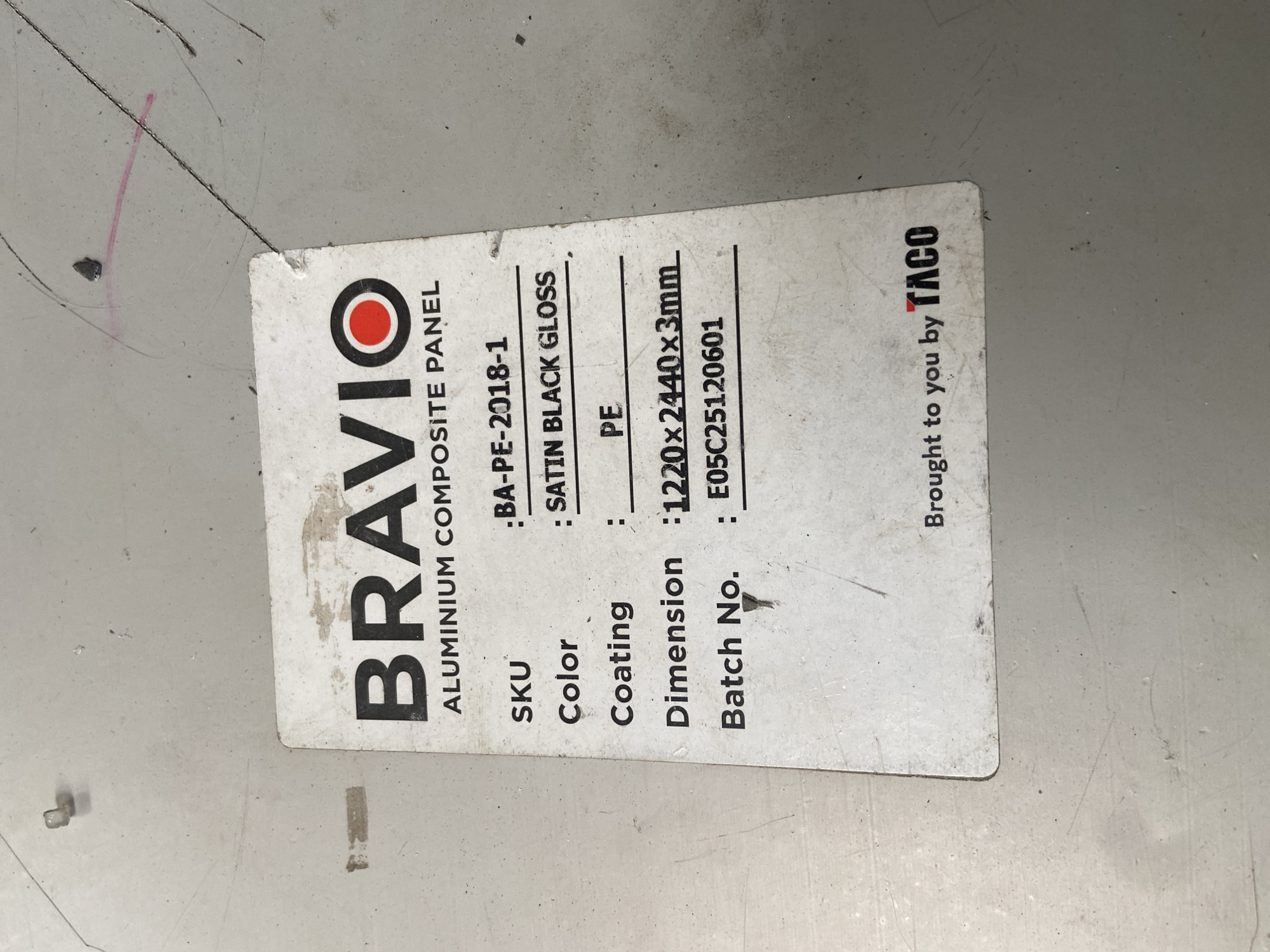 The image is a close-up, eye-level shot of a sticker on a light-colored, textured surface, likely a material panel. The sticker, which is white with black text and a red circle logo, is slightly worn and creased. It has a header that reads "BRAVIO ALUMINIUM COMPOSITE PANEL". Below this, it lists several details: "SKU : BA-PE-2018-1", "Color : SATIN BLACK GLOSS", "Coating : PE", "Dimension : 1220x2440x3mm", and "Batch No. : E05C25120601". At the bottom of the sticker, it says "Brought to you by TACO". The surface around the sticker appears to be a workshop or industrial environment, with visible scratches and some small debris. There are no people or animals in the image. The location in Plosokandang, Indonesia is not depicted or suggested by any elements within the image.