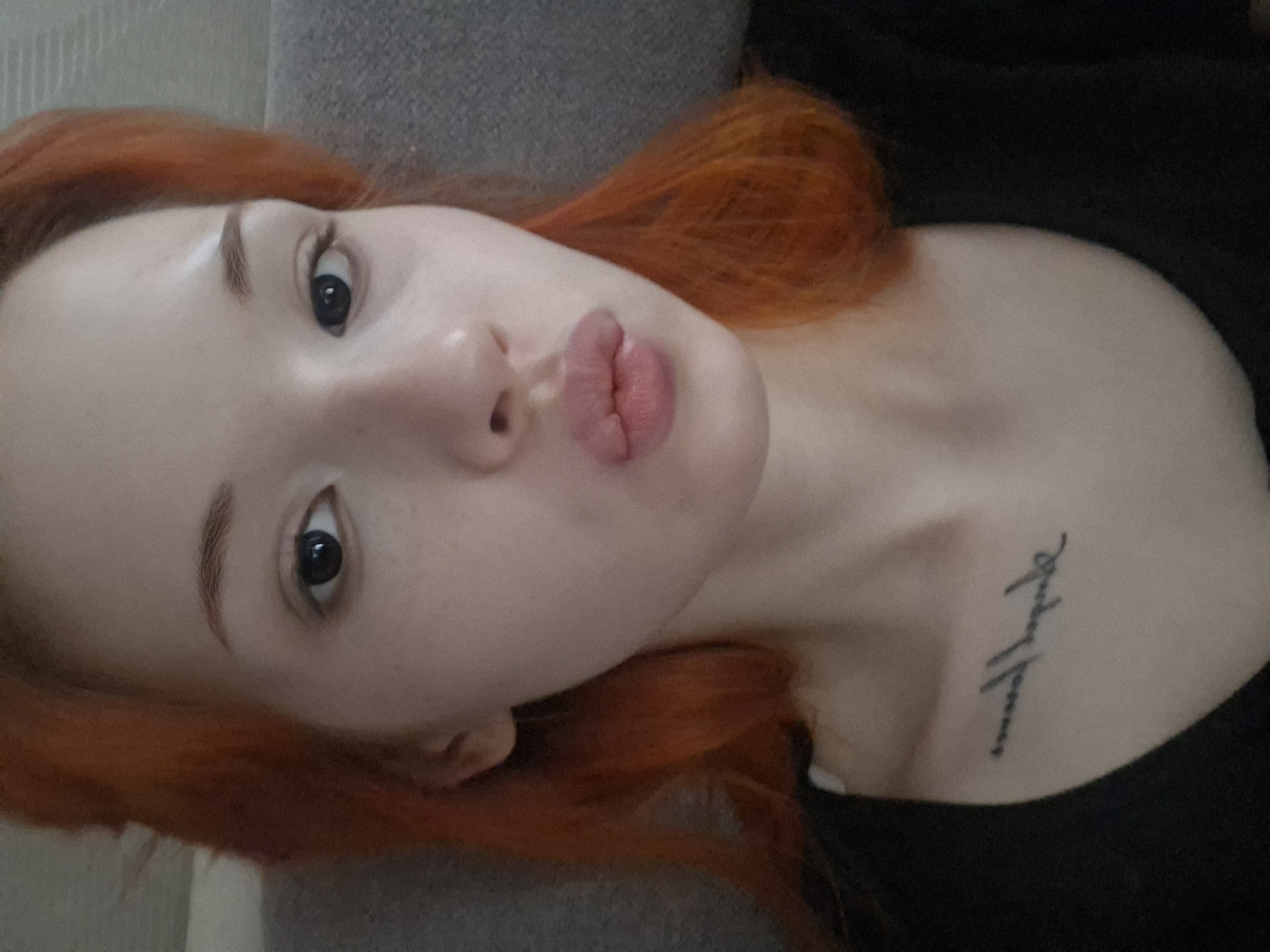 The media file presents a close-up, portrait-oriented shot of a young person, likely female, with fair skin, dark eyes, and distinctive bright red-orange hair. The subject is captured from a slightly elevated angle, looking directly into the camera with a "pout" expression, her lips pressed together.
She is wearing a dark, possibly black, garment visible around her neck and shoulder area. A small, black script tattoo is visible on her upper right chest, below the collarbone. The text of the tattoo is written in a flowing, handwritten style but is too small and stylized to be clearly deciphered.
The background is uniformly blurred and appears to be a light gray, textured fabric, suggestive of upholstery like a couch or bedding, indicating an indoor setting. The lighting is soft and even, providing no specific clues about the time of day or outdoor conditions. The overall impression is a casual, direct, and perhaps playful pose for a photograph.