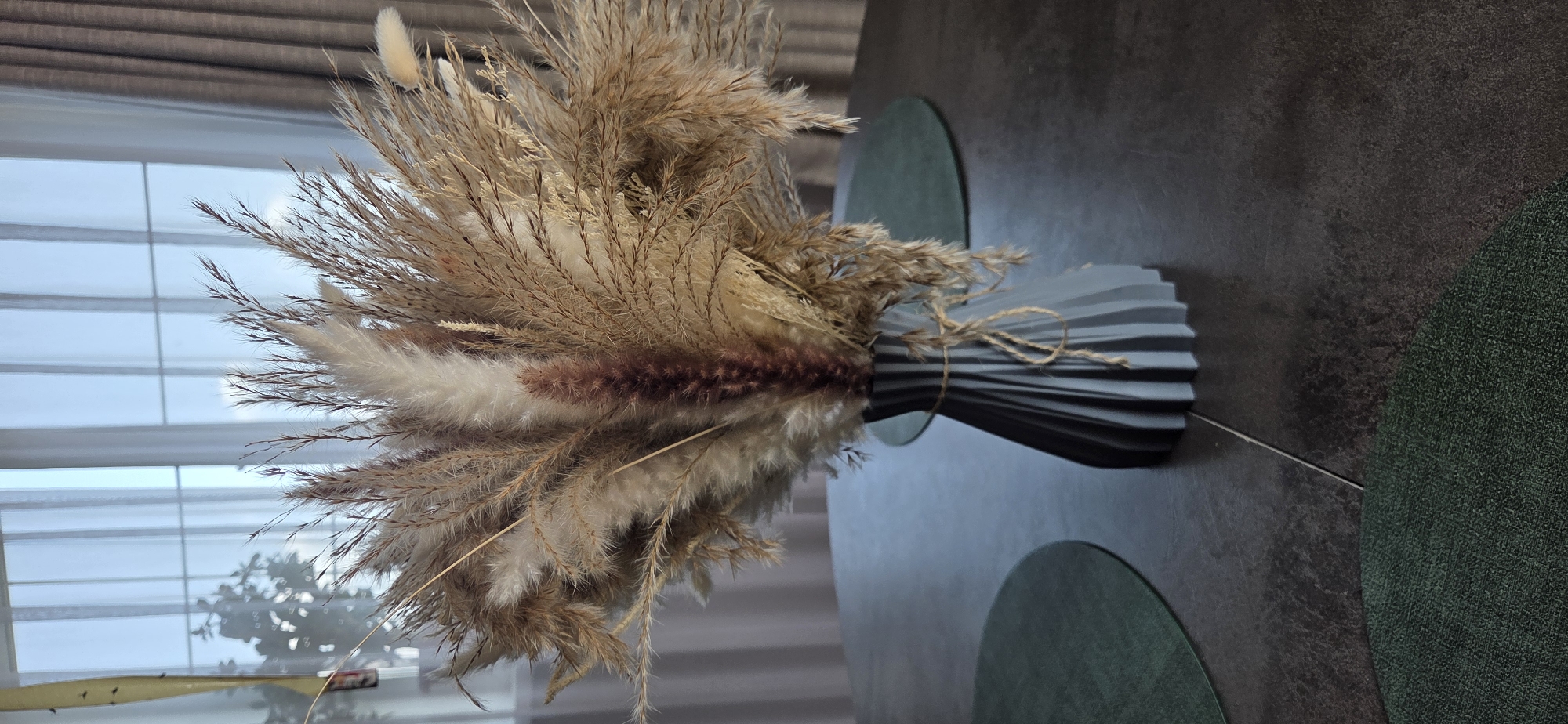 In an indoor setting, a dark grey, fluted vase stands on a dark textured table. The vase is tied with a piece of twine and contains a large bouquet of dried pampas grass and other dried floral elements. The bouquet features a variety of textures and colors, ranging from creamy white to light brown and a darker reddish-brown in the center. Behind the vase, a window is visible with sheer white curtains and darker drapes, allowing diffused light to enter the room. The window has a grid pattern that suggests it is divided into panes. Two dark green, circular placemats are partially visible on the table, one to the left and one to the right of the vase. The overall scene depicts a still life arrangement in a domestic interior. The image does not contain any elements that would suggest a specific location in the city of Маркова, Russia, beyond being an indoor space.
