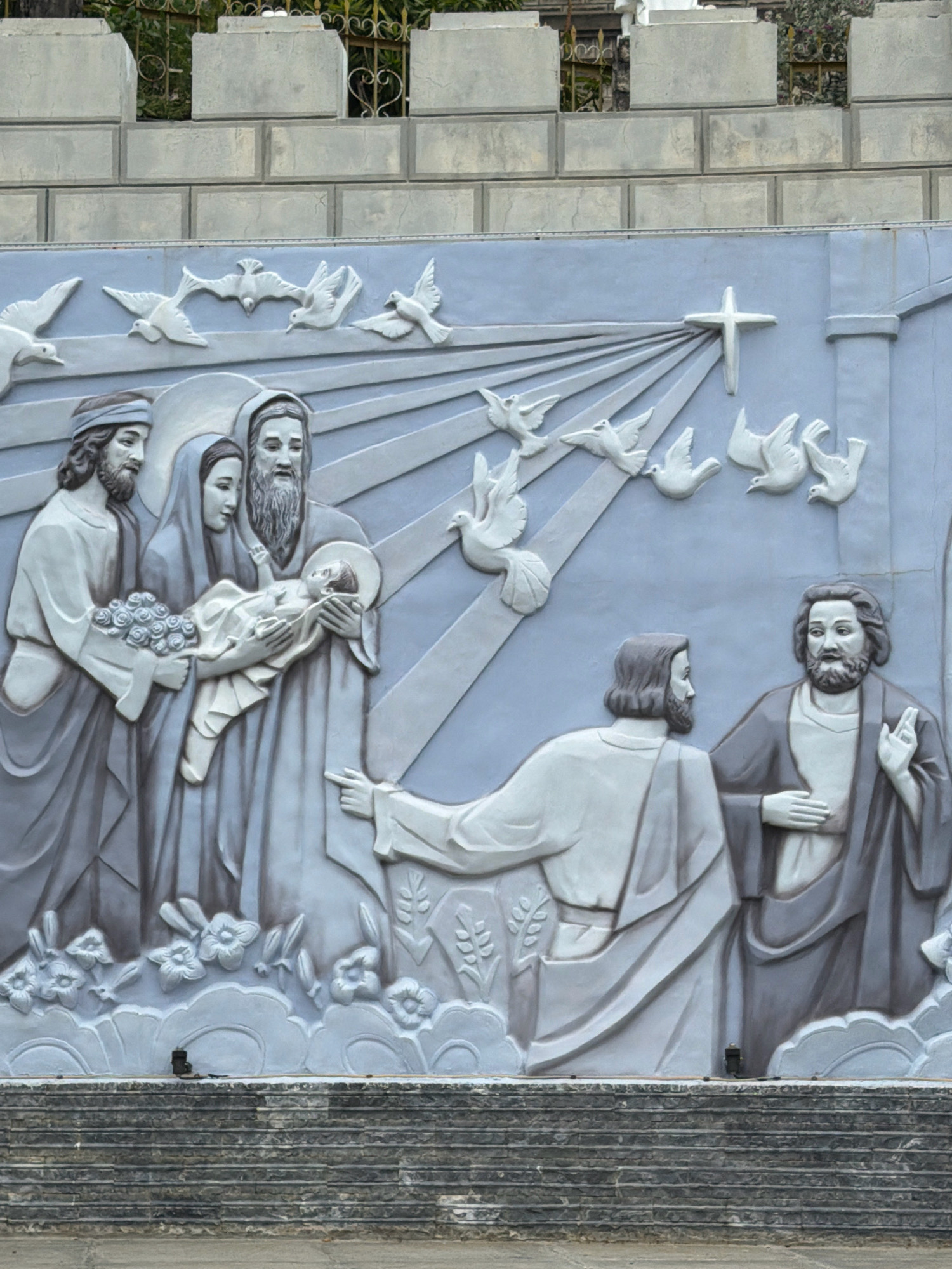 This image depicts a relief sculpture on a light blue wall, likely a monument or religious site. The sculpture is rendered in shades of grey and white, creating a sense of depth and texture.
The central focus of the sculpture is a group of figures, appearing to represent a biblical scene. On the left, a man with a beard and a headscarf holds a bouquet of flowers. Next to him, a woman with a veiled head gazes tenderly at a baby she is holding. An older man with a long beard stands behind them, also looking at the baby.
To the right of this group, two men are depicted in conversation. One man, with a beard and wearing a robe, gestures with his right hand. The other man, seen from the back, also wears a robe.
Above the figures, several white doves are sculpted in flight, some with their wings outstretched. A stylized cross is visible in the upper right portion of the sculpture, with rays of light emanating from it. Below the figures, there are sculpted flowers and clouds.
The sculpture is situated above a stone staircase made of layered grey stones. In the background, above the light blue wall, is a light grey brick wall topped with decorative stone elements. Some greenery is visible peeking through openings in the background wall.
The style of the sculpture suggests a religious theme, likely Christian. The presence of doves and a cross are strong indicators.
This relief sculpture is located at the Long Son Pagoda in Nha Trang, Vietnam. The pagoda is a prominent Buddhist temple known for its large white Buddha statue and the dragon motifs adorning its roofs. While this specific sculpture appears to have Christian iconography, it is not uncommon for religious sites in Vietnam to incorporate diverse artistic and symbolic elements. The Long Son Pagoda is situated in the Phuong Son ward of Nha Trang city.