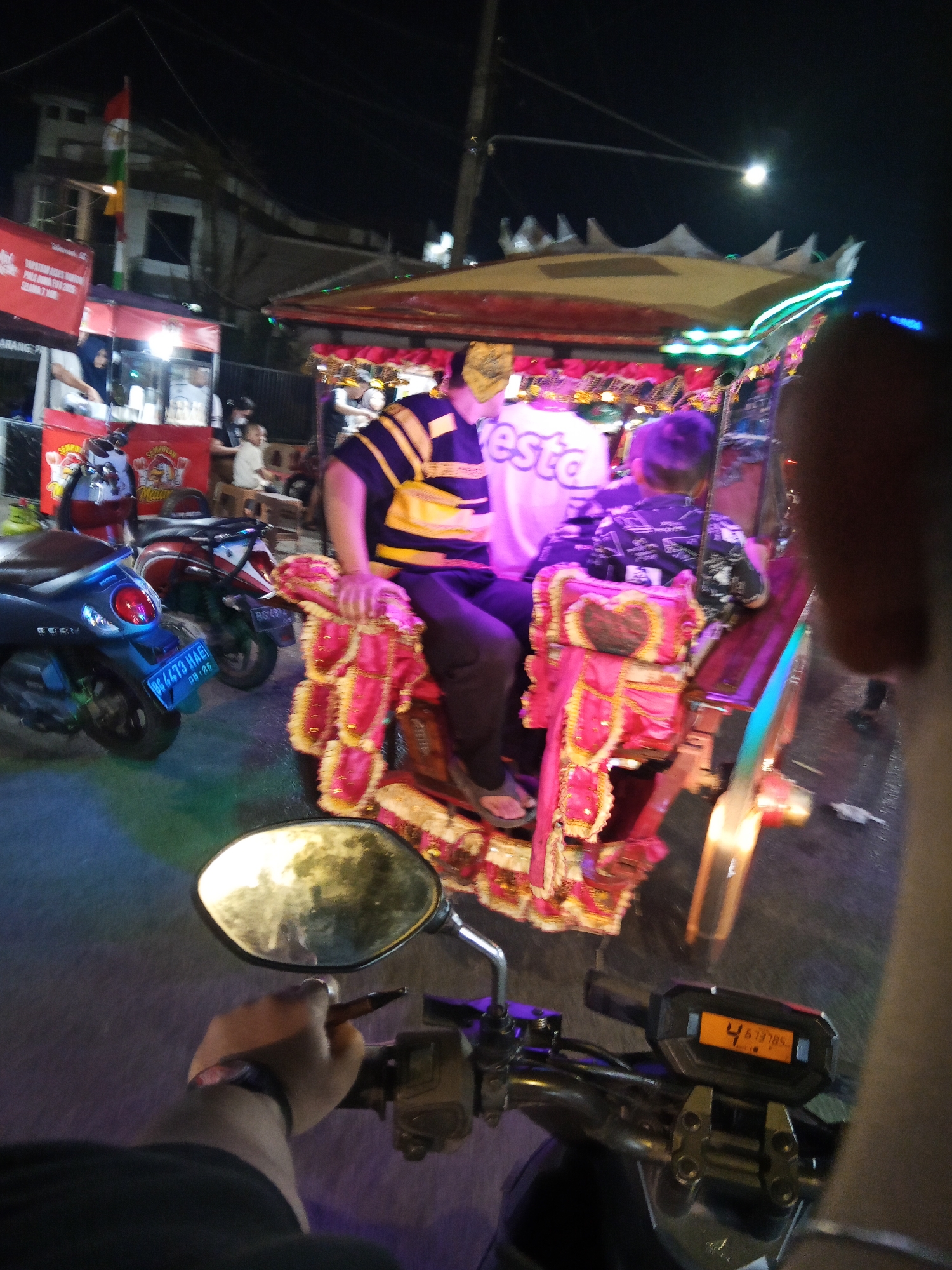The image depicts a night scene in Lubuklinggau, Indonesia, likely at a street market or festival, given the illuminated stalls and a decorated vehicle. In the foreground, a hand is visible on the handlebars of a motorcycle, holding a pen. The motorcycle's digital display shows "4 67378.5". A motorcycle with license plate "BG 4473 HAE" is parked to the left. Behind it, another red and white motorcycle is visible. Dominating the mid-ground is a brightly decorated, multi-colored vehicle, possibly a pedicab or modified auto-rickshaw, with people seated inside. One individual in the vehicle wears a yellow and black striped shirt and a mask. Further back, food stalls with red awnings and a flag are visible. The overall environment suggests a lively, outdoor evening event in an urban Indonesian setting.