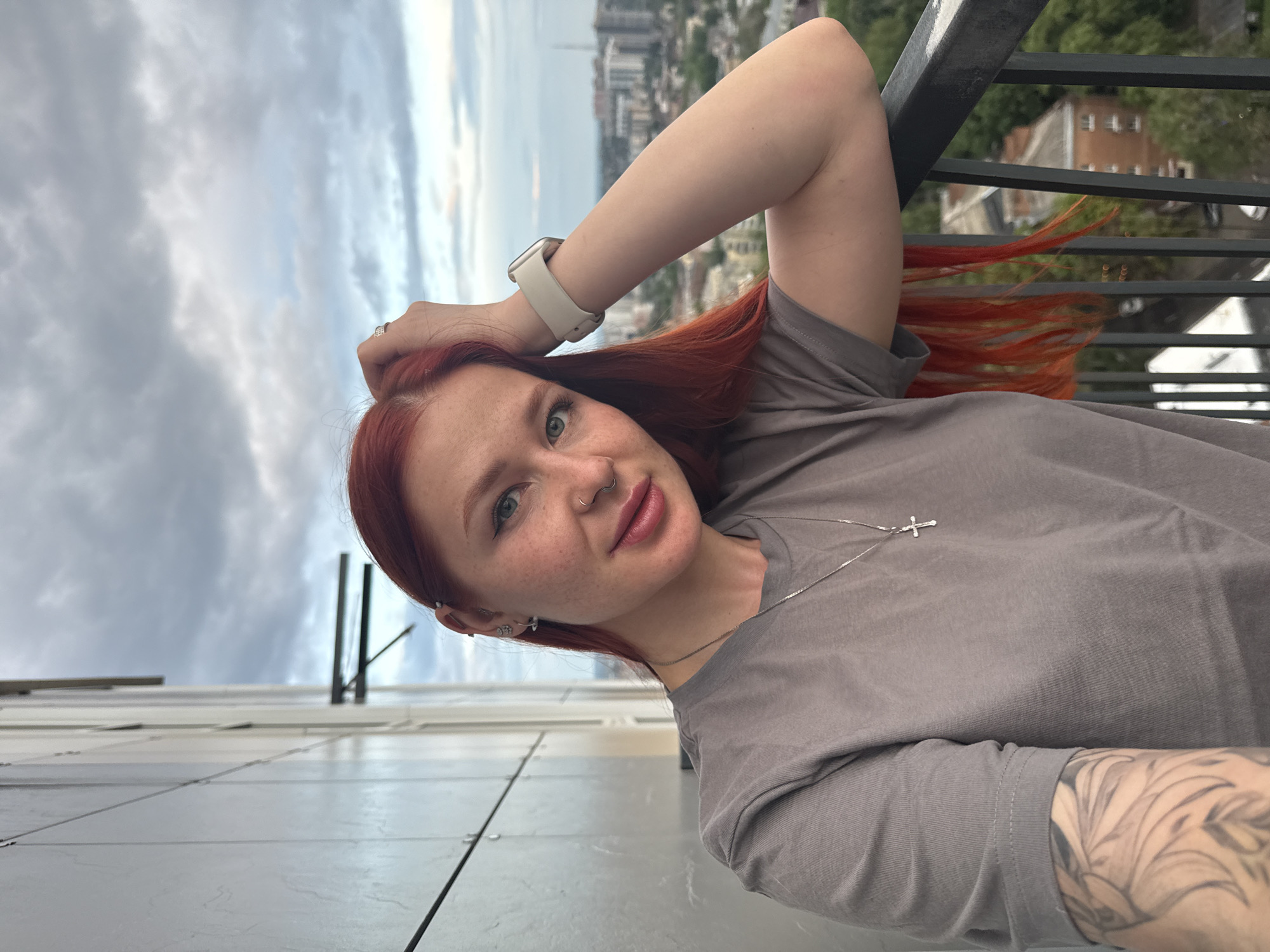 A medium shot, taken from a low angle, depicts a woman with long, red hair against an urban backdrop. She is leaning against a dark-colored metal railing, with her right arm raised and her hand near her head. She is wearing a grey t-shirt, a silver necklace with a cross pendant, and a white smartwatch on her left wrist. Tattoos are visible on her left forearm. Her facial features include freckles and a nose ring. In the background, buildings and a cloudy sky are visible, partially obscured by the railing. The location is Rostov-on-Don, Russia.