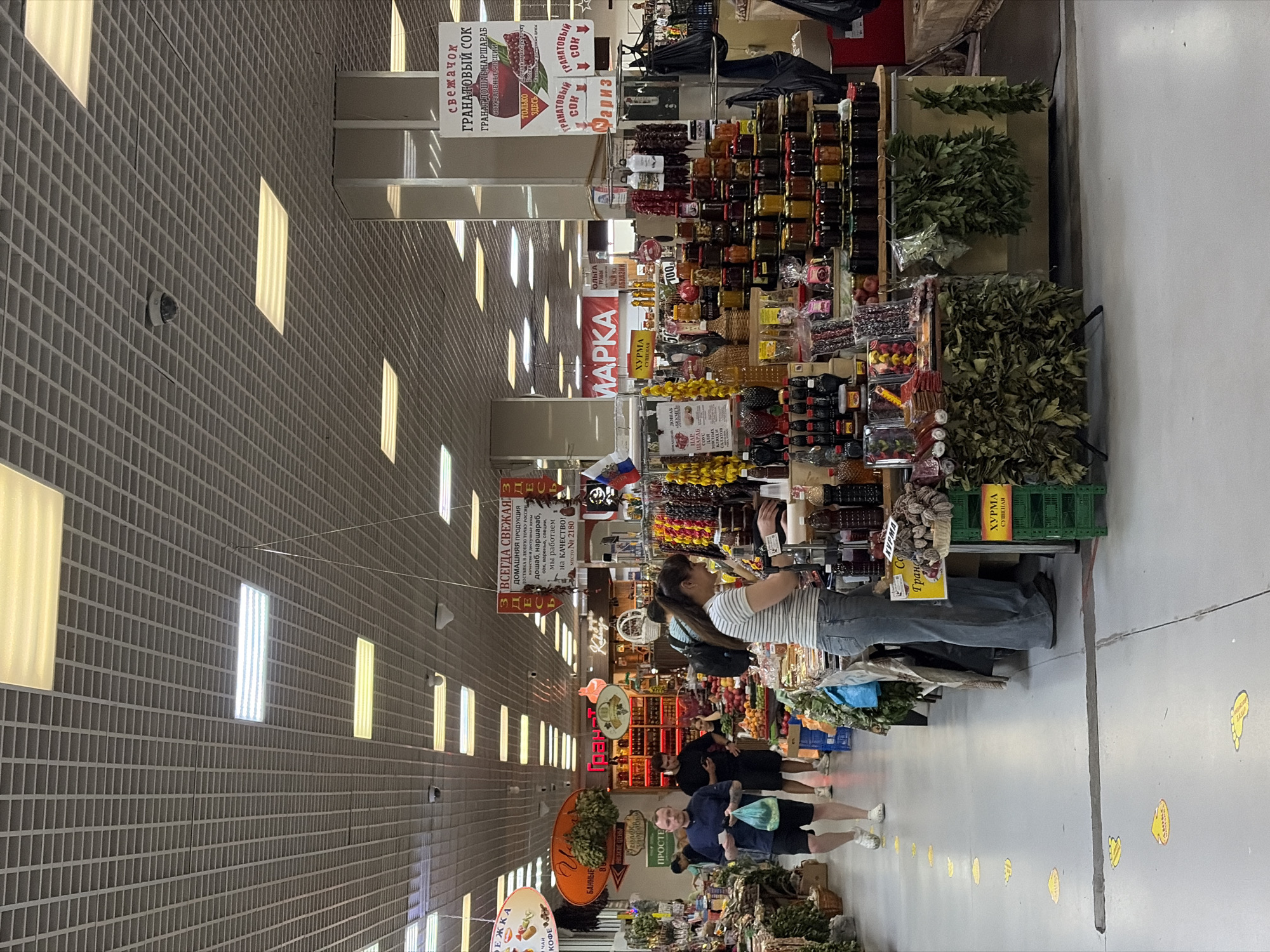 The image shows an indoor market stall in Sochi, Russia, filled with various food items. A woman in a striped shirt and jeans is standing at the stall, appearing to examine or handle products. Behind her, a display of numerous glass jars filled with colorful preserves and jams is arranged on shelves. To the left, a man in a dark shirt and shorts is walking away from the camera, carrying a green plastic bag. Another man in a black t-shirt and shorts is seen walking past him. The market ceiling is a grid of acoustic tiles with fluorescent lighting panels. Signage in Russian is visible throughout the scene, indicating product names and prices. The floor is concrete with yellow foot-shaped stickers, possibly indicating social distancing markers. The overall atmosphere suggests a bustling marketplace.