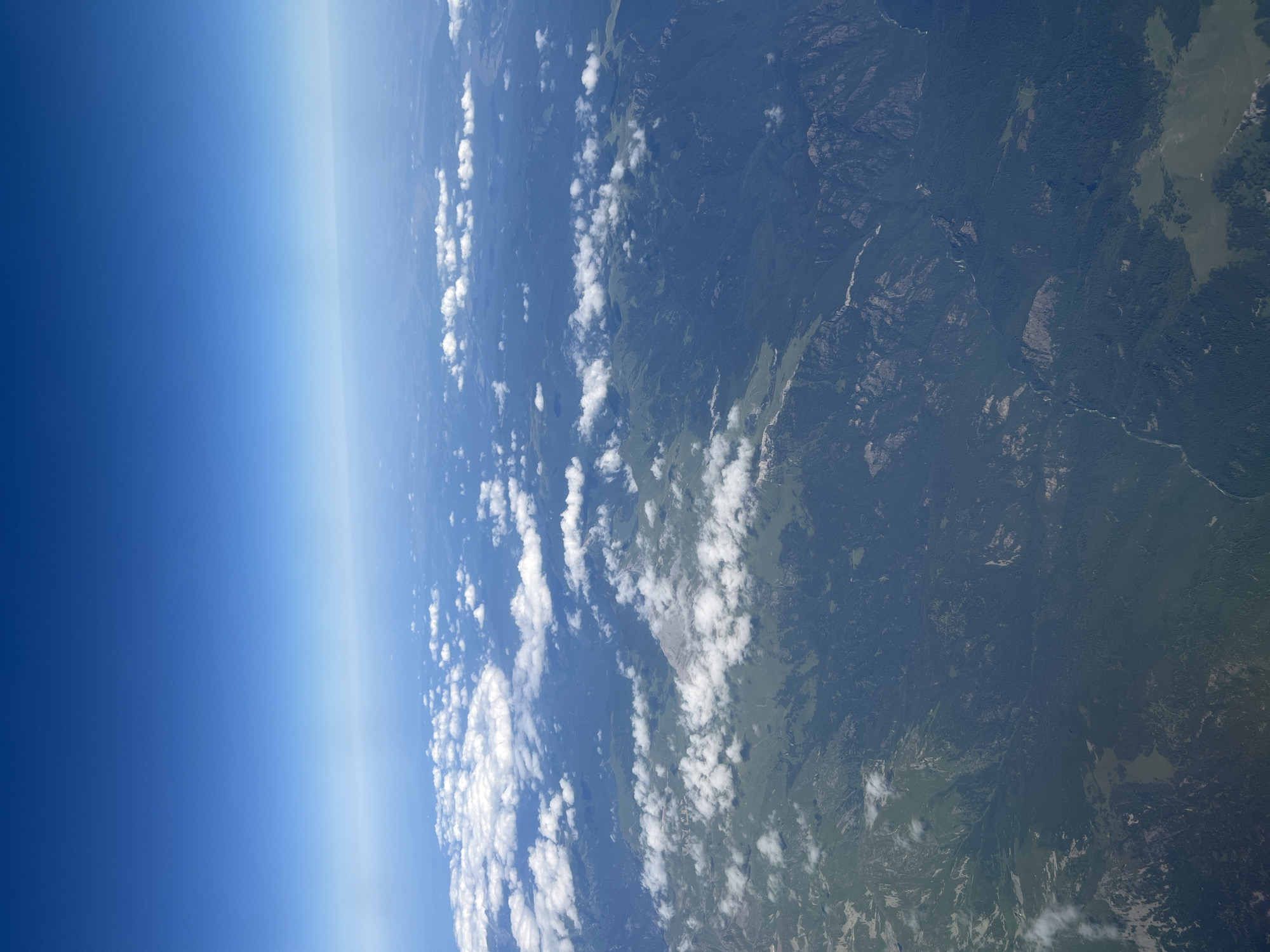 This is an aerial photograph taken from an airplane, looking down on a vast expanse of mountainous terrain covered in dense green forests. The sky is a clear, bright blue with a gradient that fades into a hazy horizon. A scattering of fluffy white clouds drifts across the landscape, casting shadows on the land below. The mountains appear rugged and uneven, with some areas showing rocky outcrops and steep slopes. Rivers or streams can be seen winding through the valleys. The overall impression is one of a remote and wild natural landscape. There is no indication of the city of Sirius, Russia, or any specific landmarks within that city.