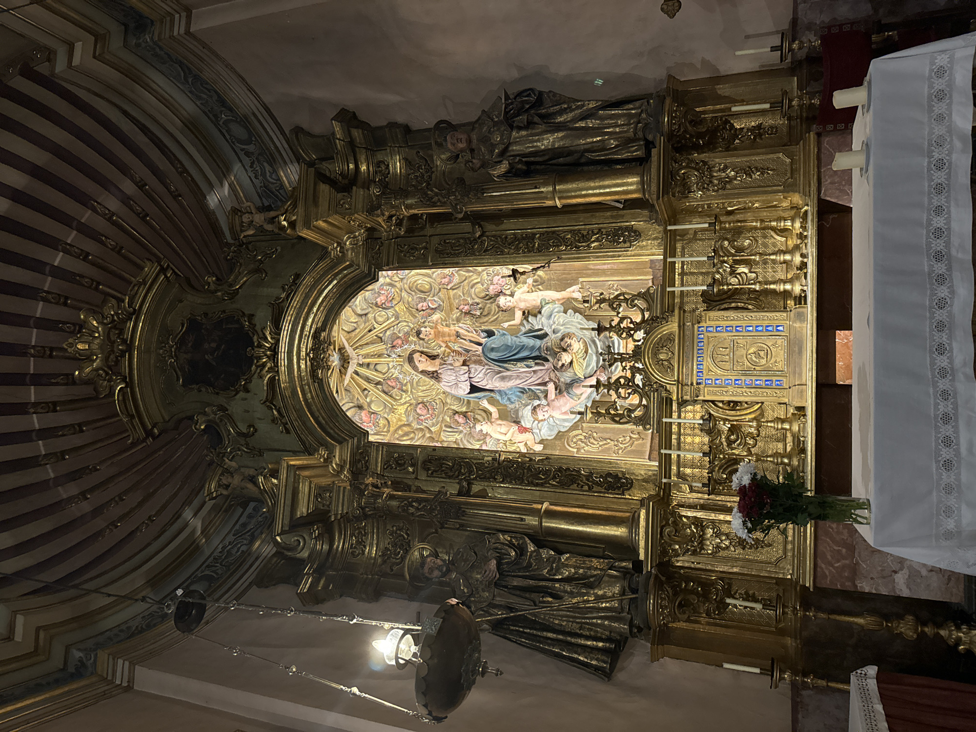 The image depicts the interior of a church altar, likely in Sóller, Spain, adorned with ornate gold detailing. The central focus is a statue of the Virgin Mary holding the infant Jesus, surrounded by cherubic figures and a dove, all illuminated by a golden glow. Flanking the central tableau are two larger statues of robed male saints, one on each side. The altar itself is covered with a white lace-trimmed cloth, set before a gilded retable featuring multiple candleholders and a decorative panel. A vase of flowers, with red and white blooms, sits on the altar. The church's ceiling is visible at the top, featuring painted stripes and decorative moldings. The overall impression is one of solemnity and religious devotion.