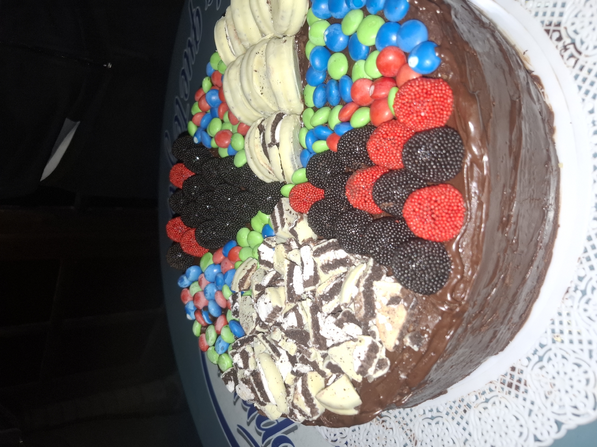 The image shows a chocolate cake decorated with candies. The cake is round with chocolate frosting. The top is divided into sections, each decorated with a different type of candy: M&Ms (red, blue, green), red and black sprinkle-covered candies, white chocolate slices, and cookies and cream pieces. The cake is on a white decorative paper doily, which is on a blue surface. The background is dark. The scene appears to be indoors. There is no indication that the image was taken in Buenos Aires, Argentina.