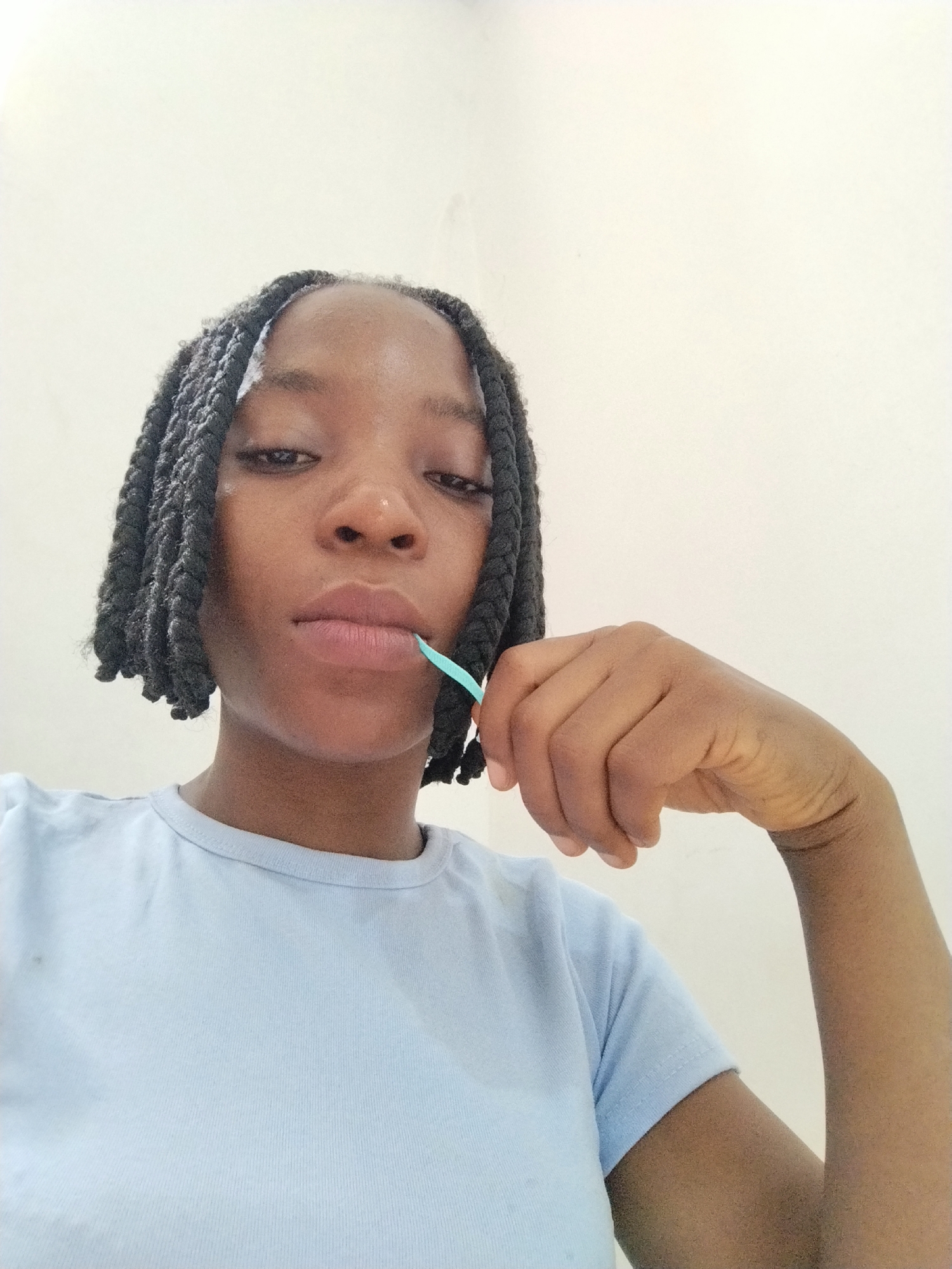 A close-up, waist-up shot shows a young Black woman from Uselu, Nigeria. She has her hair braided into medium-sized box braids that fall to her shoulders. She is wearing a light blue t-shirt. Her right hand is raised to her face, and she is holding a thin, light blue object, possibly a piece of floss, between her lips. The background is a plain, off-white wall.