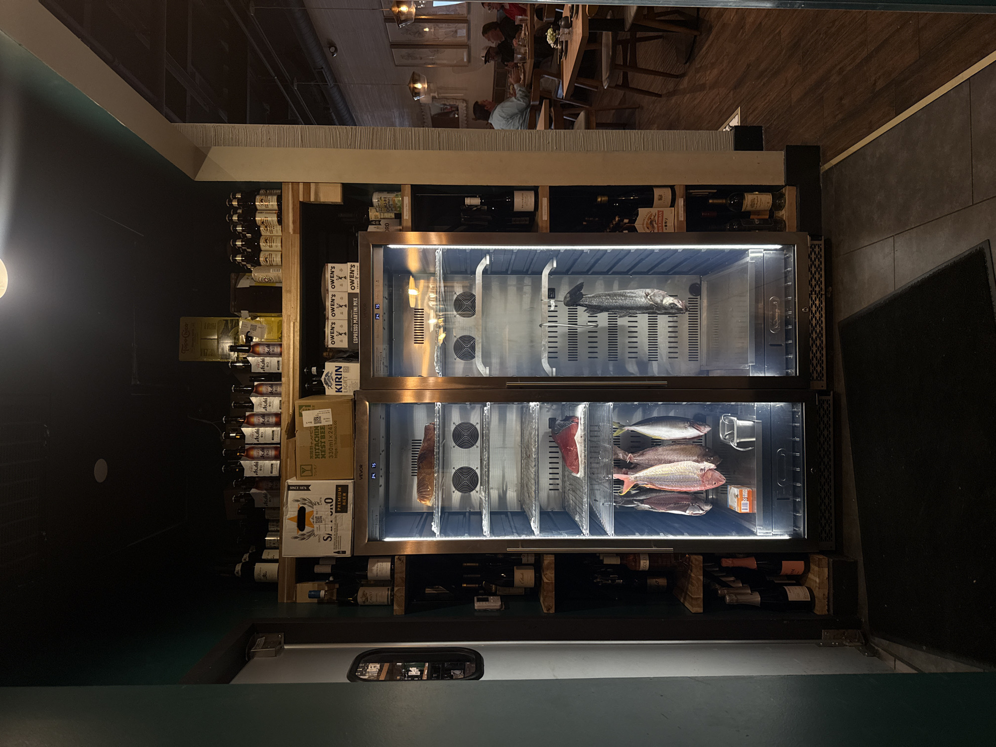The image shows an interior scene, likely a restaurant in Southlake, United States of America. The primary focus is a two-door refrigerated display case showcasing a variety of fish on separate racks, with built-in fans. The display case is surrounded by shelves stocked with bottles of wine and beer. Brands of beverages visible include Topo Chico, Asahi, Hitachino Nest Beer, Kirin Light, Owen's Espresso Martini Mix, and Soto Premium Beer. The shelves are set against a dark-colored wall. In the background, a blurred view of people seated at tables suggests a dining area. The floor is tiled and covered by a black mat. The lighting is a mix of natural and artificial, with reflections visible on the glass doors of the display case.
