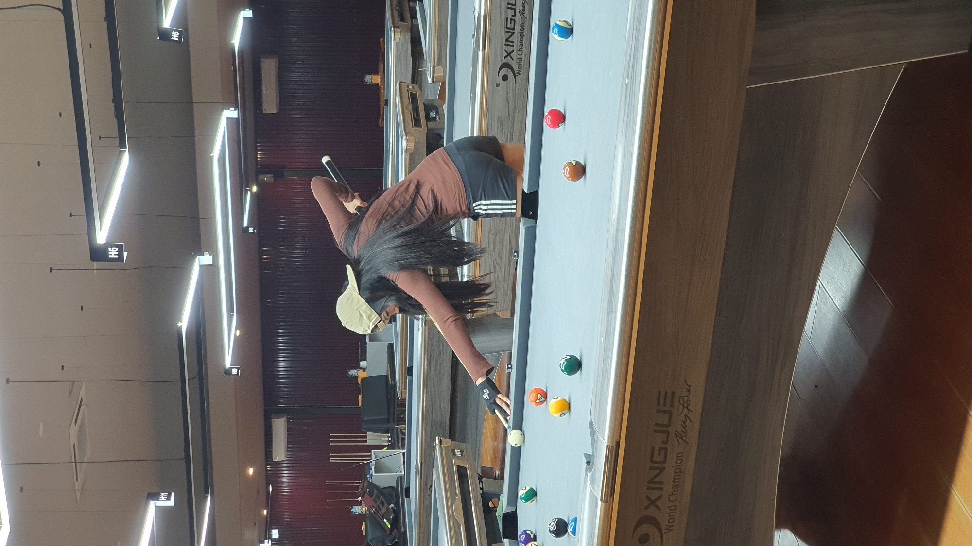 A young woman with long dark hair and a tan baseball cap is playing billiards in an indoor pool hall. She is wearing a brown long-sleeved shirt and black shorts with white stripes. She is in the process of striking a white cue ball with a black pool cue, aiming for other balls on the table. Several colorful billiard balls are scattered across the grey felt surface of the pool table. The environment appears to be a modern facility with multiple pool tables, track lighting on the ceiling, and dark-colored walls. The image also shows signage on the pool table that reads "XINGJUE World Champion". The location is identified as Kuta Selatan, Indonesia.