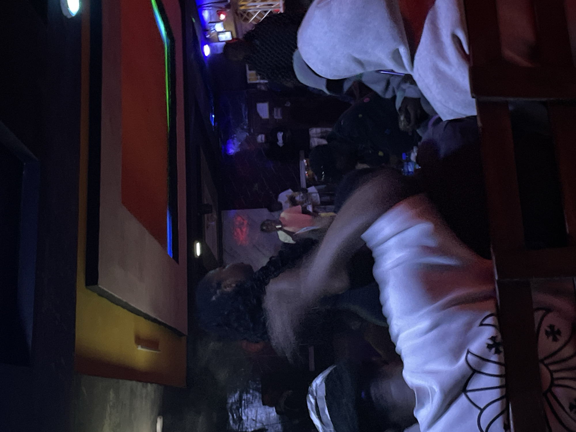 In Durumi, Nigeria, a dimly lit interior scene captures a group of individuals gathered in what appears to be a nightclub or a similar entertainment venue. The lighting is moody, with colorful neon lights and spotlights illuminating the space, casting shadows and highlighting the subjects.
In the foreground, a person wearing a white hoodie with a distinct black graphic design on the sleeve is seated, their back mostly to the camera. To their left, another individual with a dark shirt and a black and white patterned cap is partially visible, also seated.
Further into the venue, a woman with dark, curly hair is standing and appears to be interacting with someone. She is wearing a black top. To her right, another person in a light-colored t-shirt is standing and holding a microphone, suggesting they might be a performer or a host.
Other individuals are visible in the background, some seated, some standing, creating a sense of a social gathering. The environment features a stage-like area in the background with lighting equipment. The ceiling has a modern, multi-layered design with integrated lighting. The overall atmosphere is one of an active social event in a public indoor space.