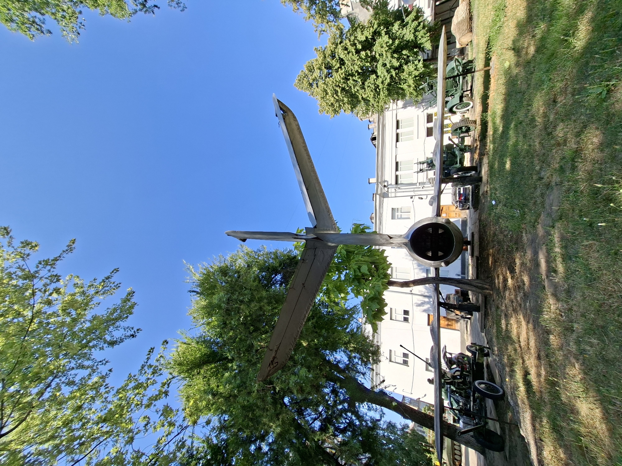 The image depicts an outdoor exhibit, likely in a museum or memorial park, featuring historical military equipment under a clear blue sky. The central and most prominent object is the tail section of a jet aircraft, viewed from the rear. Its twin wings extend outwards, angled upwards. Beneath the aircraft's tail, a row of wheeled military artillery pieces are displayed. To the left, a dark-colored artillery piece with a machine gun mounted on it is visible. Further to the right, a lighter green artillery piece is positioned, and behind it, another similar green artillery piece is partially obscured. A white building with multiple windows forms the backdrop, partially covered by the lush green foliage of trees. The ground is covered with grass and dry leaves, suggesting a park-like setting. The scene is illuminated by bright sunlight, casting shadows on the grass. The image is taken from a low angle, looking up towards the sky and the aircraft. The specific location is the Museum of Military History in Chișinău, Moldova. There are no people visible in the image.