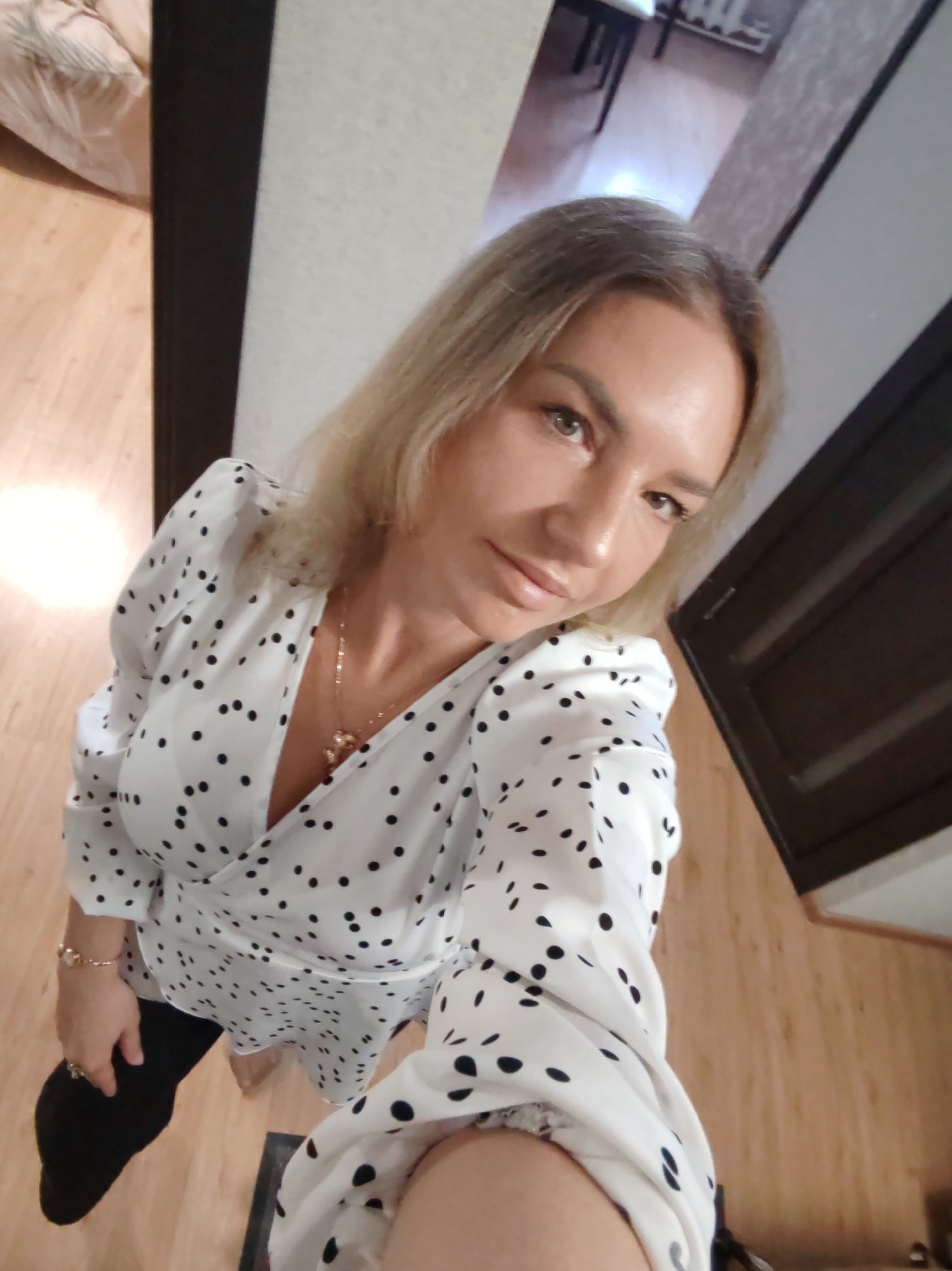 A woman is captured in a high-angle, full-body selfie, looking directly at the camera with a calm, neutral expression. She has blonde to light brown hair and appears to be of fair complexion, with light brown eyes. She is wearing a long-sleeved, white wrap-style dress adorned with numerous black polka dots, featuring a V-neckline and puffed sleeves. A gold-colored necklace with a distinctive, multi-element pendant, possibly depicting stylized crosses or flowers, hangs around her neck. A gold-toned bracelet with pearl-like beads is visible on her left wrist. Black leggings or trousers can be seen beneath the dress.
The setting is indoors, likely a residential space in Petropavlovsk-Kamchatsky, Russia, characterized by a light wooden floor and dark vertical panels, possibly door frames, on either side of the frame. In the background to the left, part of a bed or sofa with light-colored bedding is visible. To the right, indistinct furniture or architectural elements can be discerned further back. The lighting suggests an indoor environment, making the specific time of day indistinguishable. There is no visible text in the image.