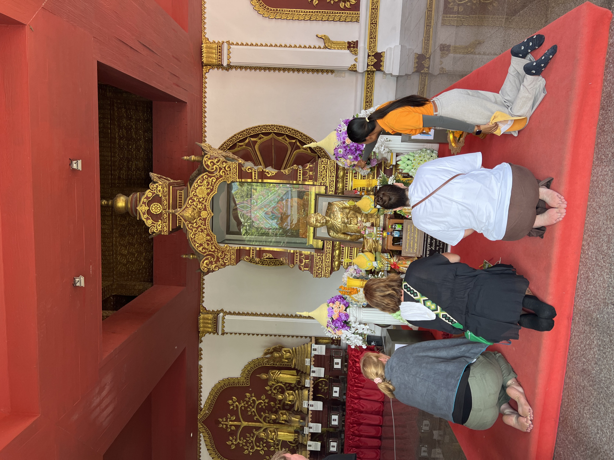 Four individuals are kneeling on a red carpet in front of a golden statue of a Buddhist monk, located indoors. The statue is housed within an ornate, gilded shrine that is decorated with intricate carvings and floral arrangements. Behind the shrine, a wall is adorned with golden Buddhist imagery and several smaller golden Buddha statues. The ceiling of the structure is painted red with exposed wooden beams. The overall scene suggests a place of religious worship, likely a Buddhist temple in Baan Hua Thanon, Thailand. The individuals appear to be engaged in prayer or respectful contemplation. One person is wearing an orange shirt, and another has a green bag slung over their shoulder. The flooring in the background is polished and reflective.