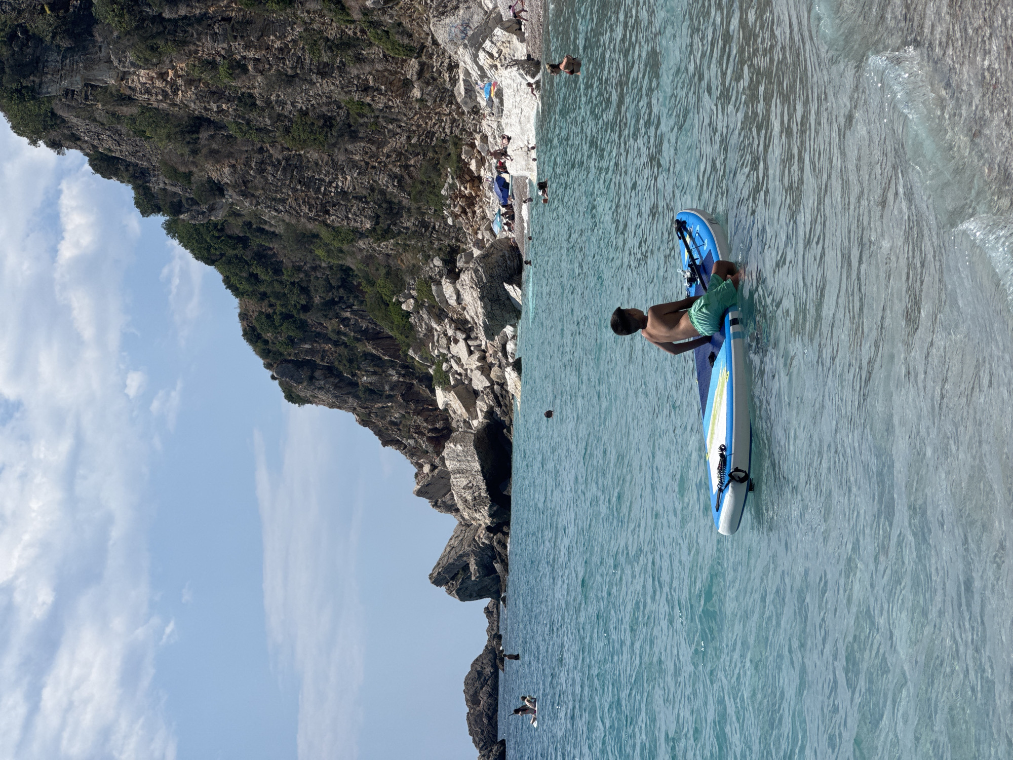 The image depicts a sunny day at a rocky beach in Mirište, Montenegro. The scene is split vertically between a rugged cliff face covered in greenery and a clear, turquoise sea. Several people are visible on the pebble beach, some sunbathing and others near the water's edge. The water is calm with gentle waves lapping at the shore.
In the foreground, a young person is kneeling on a paddleboard, which is partially submerged in the water. They are wearing green swim trunks. The paddleboard is blue and white with some black accents. The activity appears to be paddleboarding or preparing to paddleboard in the calm sea.
Further in the water, a few other individuals can be seen, some swimming or wading. In the distance, more people are scattered along the beach. The sky is a bright blue with scattered white clouds, suggesting pleasant weather. The rocky cliffs create a dramatic backdrop to the beach and sea. The overall atmosphere is one of a relaxed summer day at the seaside. No visible text is present in the image.