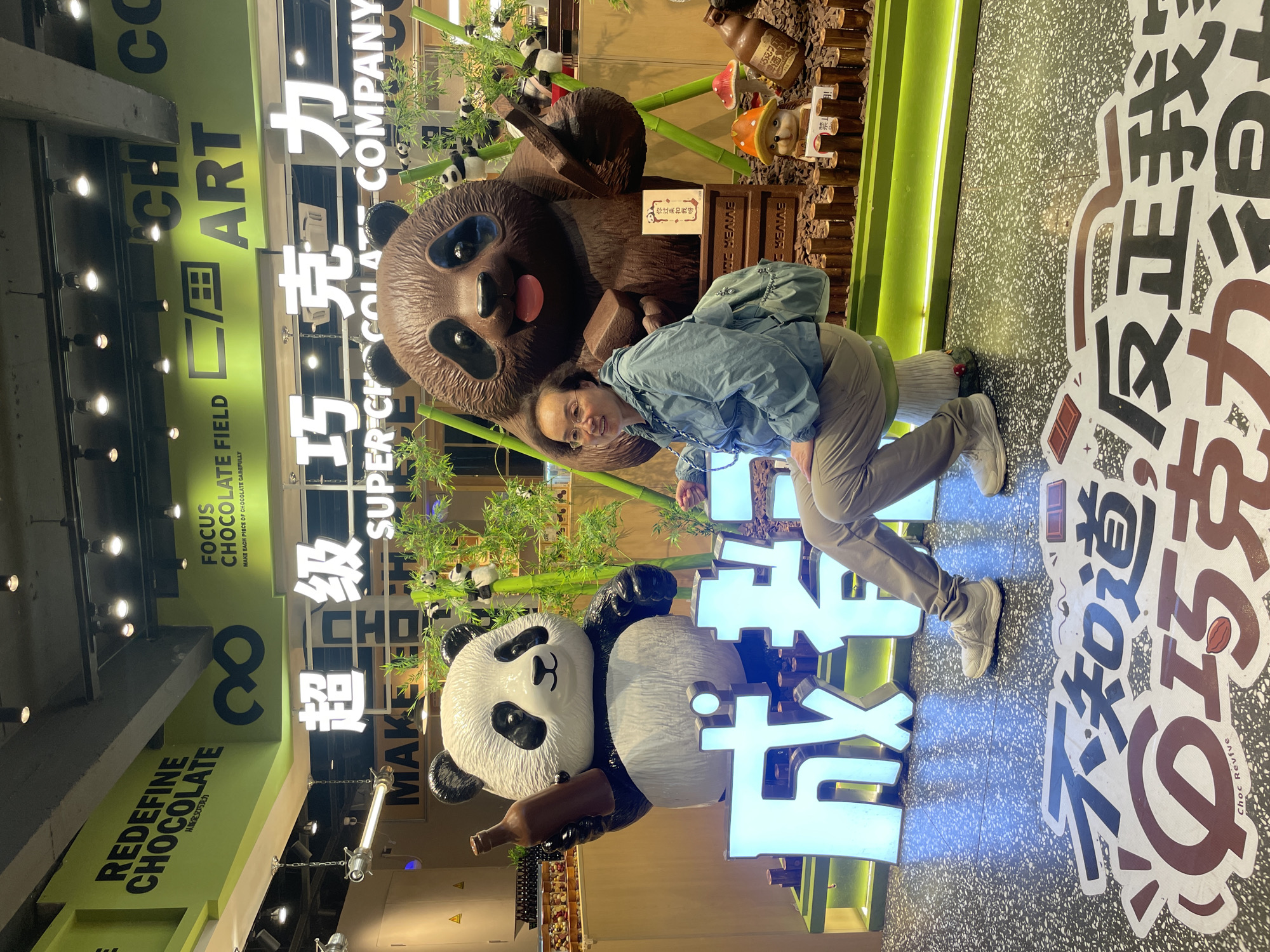 A woman in a blue jacket and khaki pants is posing next to a chocolate panda statue in what appears to be a chocolate store. The store is located in Wuhou District, China.
The woman is smiling, with her left arm around the panda. She is positioned in front of a panda statue, next to light-up Chinese characters. A large chocolate panda is visible in the background, along with bamboo and other decorative elements. Store signage is present, with text including "CHOCOLATE FIELD," "REDEFINE CHOCOLATE," and Chinese characters. The flooring has large Chinese characters on it. Overhead lighting is visible.