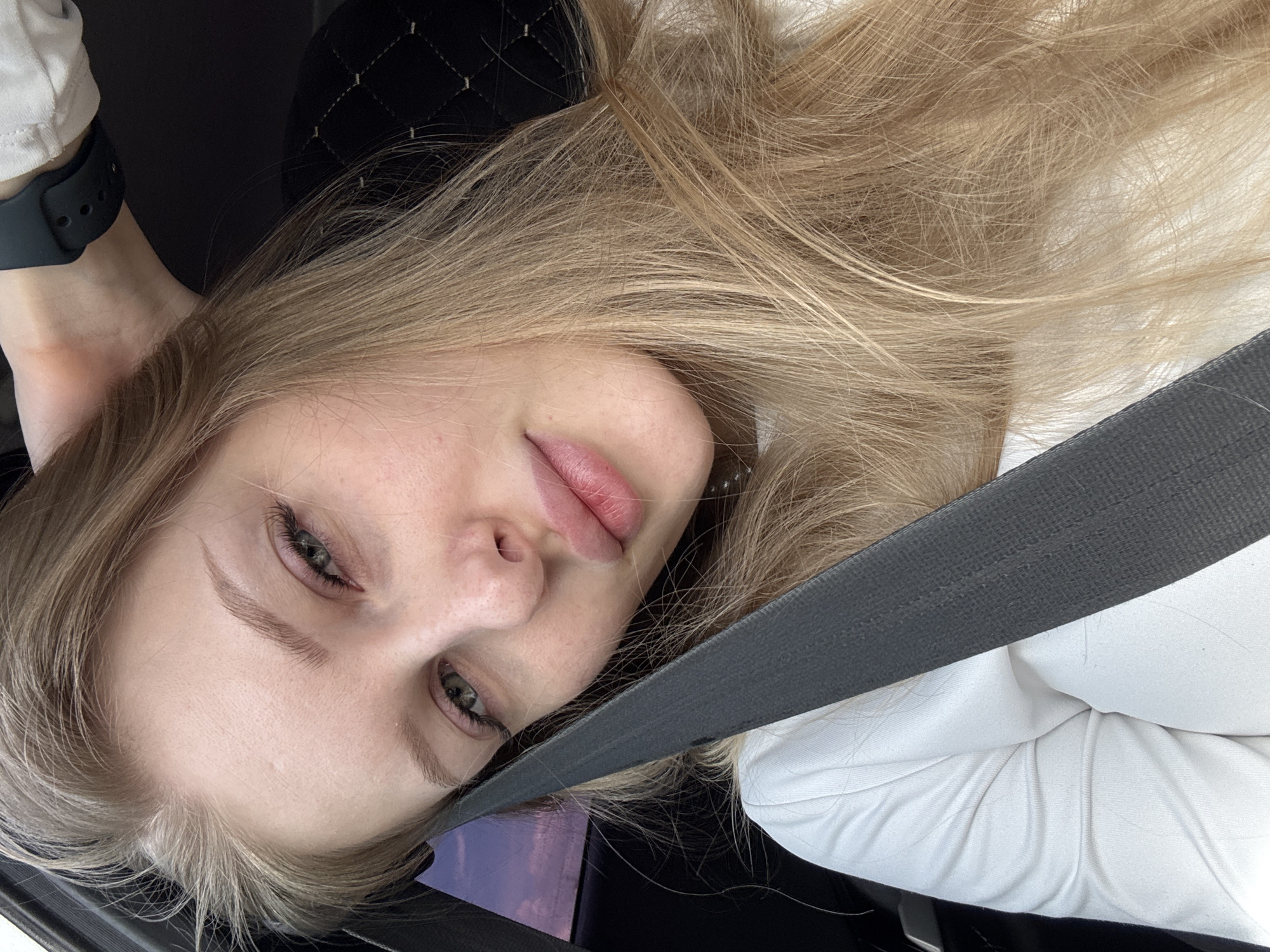 A close-up selfie captures a young woman with long, light blonde hair and blue-green eyes, appearing to be taken inside a vehicle in Perm, Russia. The image is significantly tilted, with the woman's head oriented diagonally from the top left to the bottom right of the frame, giving a casual, almost reclining posture. She gazes directly at the camera with a neutral, slightly pensive expression.
Her fair skin shows minimal makeup, highlighting her eyelashes with mascara and her well-defined, light brown eyebrows. Her lips are full and tinted with a soft pink color. Her light blonde hair, somewhat tousled, cascades around her face and over her shoulders.
She is wearing a white top or blouse, visible on her right shoulder and arm. A dark gray seatbelt diagonally crosses her chest and shoulder, indicating she is seated in a car. On her left wrist, a dark-colored smartwatch with a textured band is clearly visible. Behind her head, a dark car seat or headrest with a quilted pattern is discernible. Through a window in the background, a soft, purplish-pink sky is faintly visible, suggesting either dawn or dusk. There is no visible text in the image.