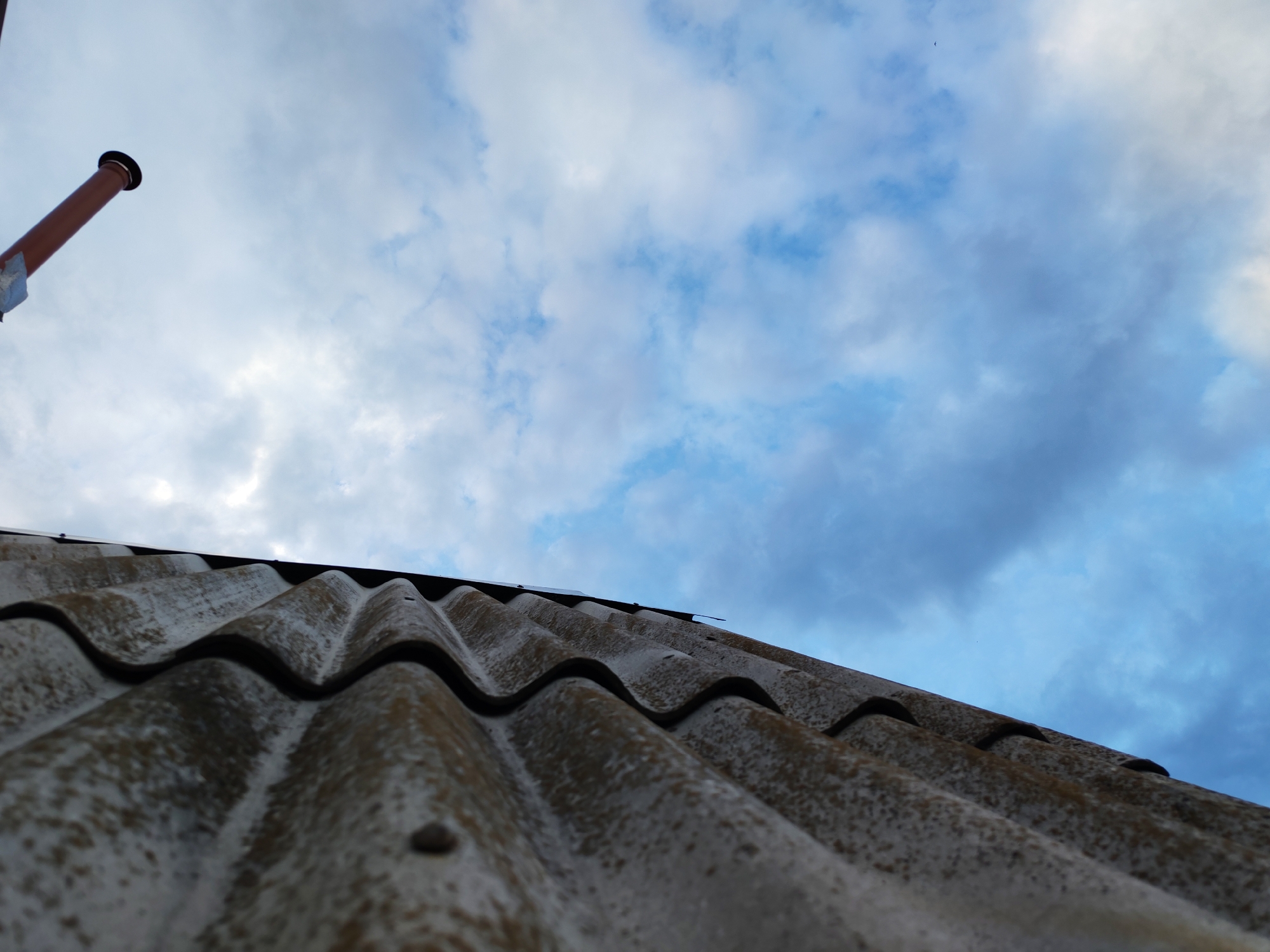 The image shows a low-angle view of a corrugated roof made of asbestos cement sheets, with a rusty metal pipe protruding from the upper left corner, likely a chimney or vent. The roof is weathered and shows signs of age, with a mottled grey and brown surface. Above the roof, a cloudy sky fills the frame, with patches of blue visible through the white and grey clouds. The lighting suggests it is either dawn or dusk, with soft, diffused light. The image appears to have been taken outdoors in the city of Pervomaysky, Russia, focusing on architectural elements and the sky. There are no people or animals visible in the scene.