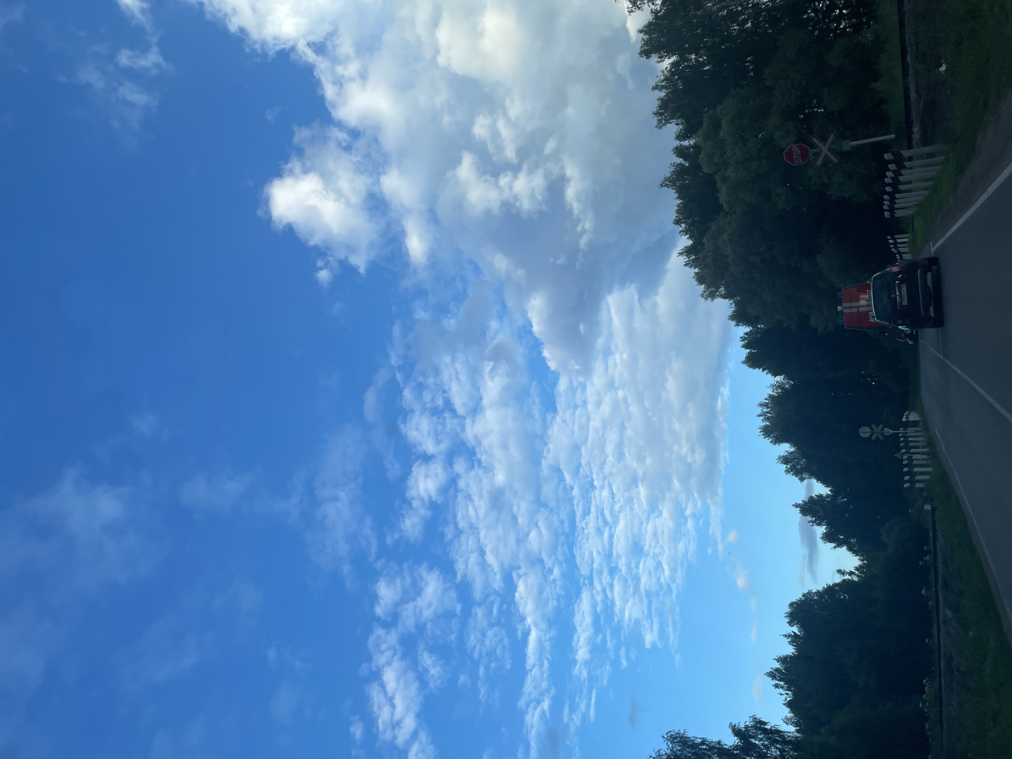The image depicts a road with trees on the side, under a blue sky with scattered clouds. A red and white vehicle, possibly a fire truck, is visible on the road, along with a railroad crossing sign. The image appears to be taken from a moving vehicle, as suggested by the slight blur and the perspective. The time of day seems to be late afternoon or early evening, given the lighting conditions and the presence of clouds. The overall mood is one of travel and observation, with a sense of natural beauty due to the sky and trees. The text "СТОП" is visible on a red octagonal sign, which is a stop sign. The license plate of the vehicle is also visible, but the text is not legible. The location is identified as Kamkoye, Russia.