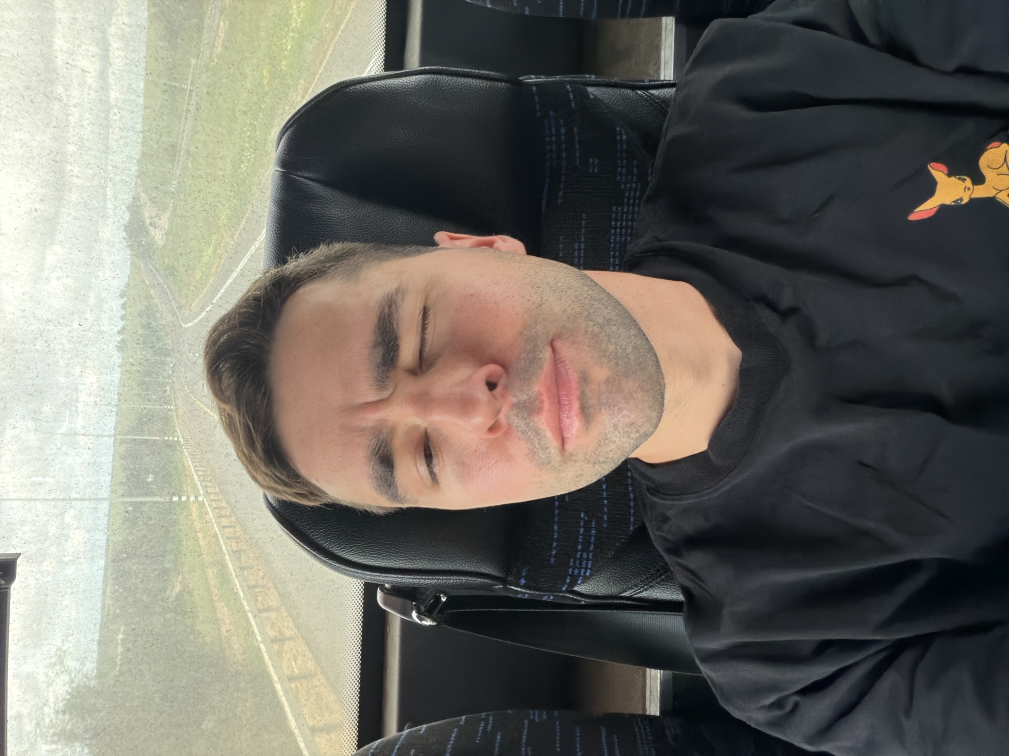 An adult male is depicted in a close-up, seated inside a bus in the Shafir Regional Council, Israel. He has short brown hair, light skin, and stubble, and is wearing a black long-sleeved shirt with an orange-eared cartoon deer emblem on the right chest. His right eye is closed, and his left eye is partially open, with a neutral mouth expression. A black seatbelt is visible over his left shoulder. The background, seen through a speckled window, shows a two-lane asphalt road with a metal guardrail, flanked by green grass and distant foliage under a cloudy sky. Several utility poles are visible along the roadside.