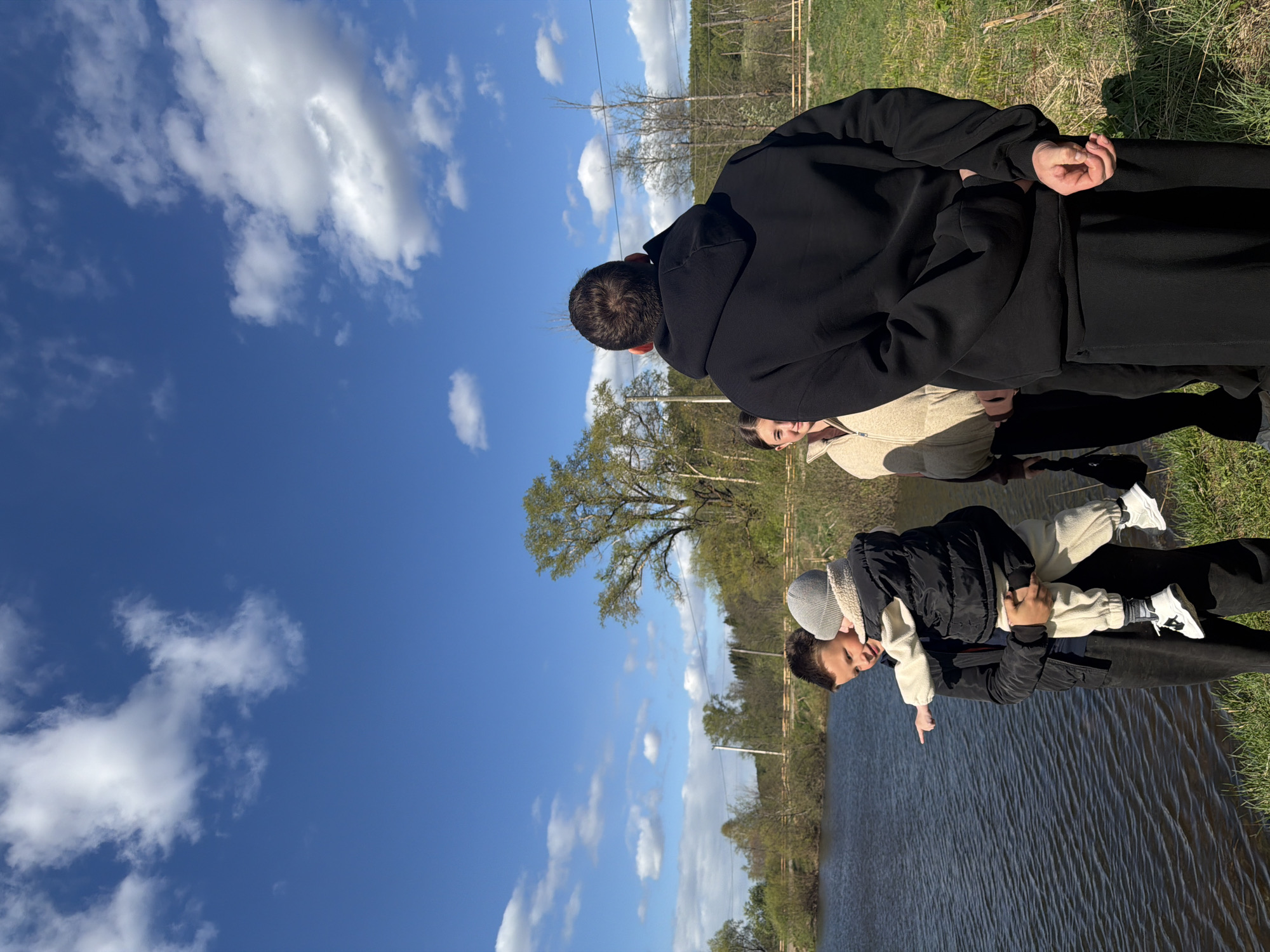 The image depicts three individuals on the bank of a body of water, likely a lake or river, in Bolshoy Bukor, Russia. One individual, wearing a black jacket and holding a young child dressed in a grey hat, black puffer jacket, and white pants, is pointing towards the water. A second individual, wearing a tan jacket, stands next to them, and a third, wearing a black hooded sweatshirt, stands behind them. The sky is blue with scattered clouds. The environment includes trees and grass along the shoreline.