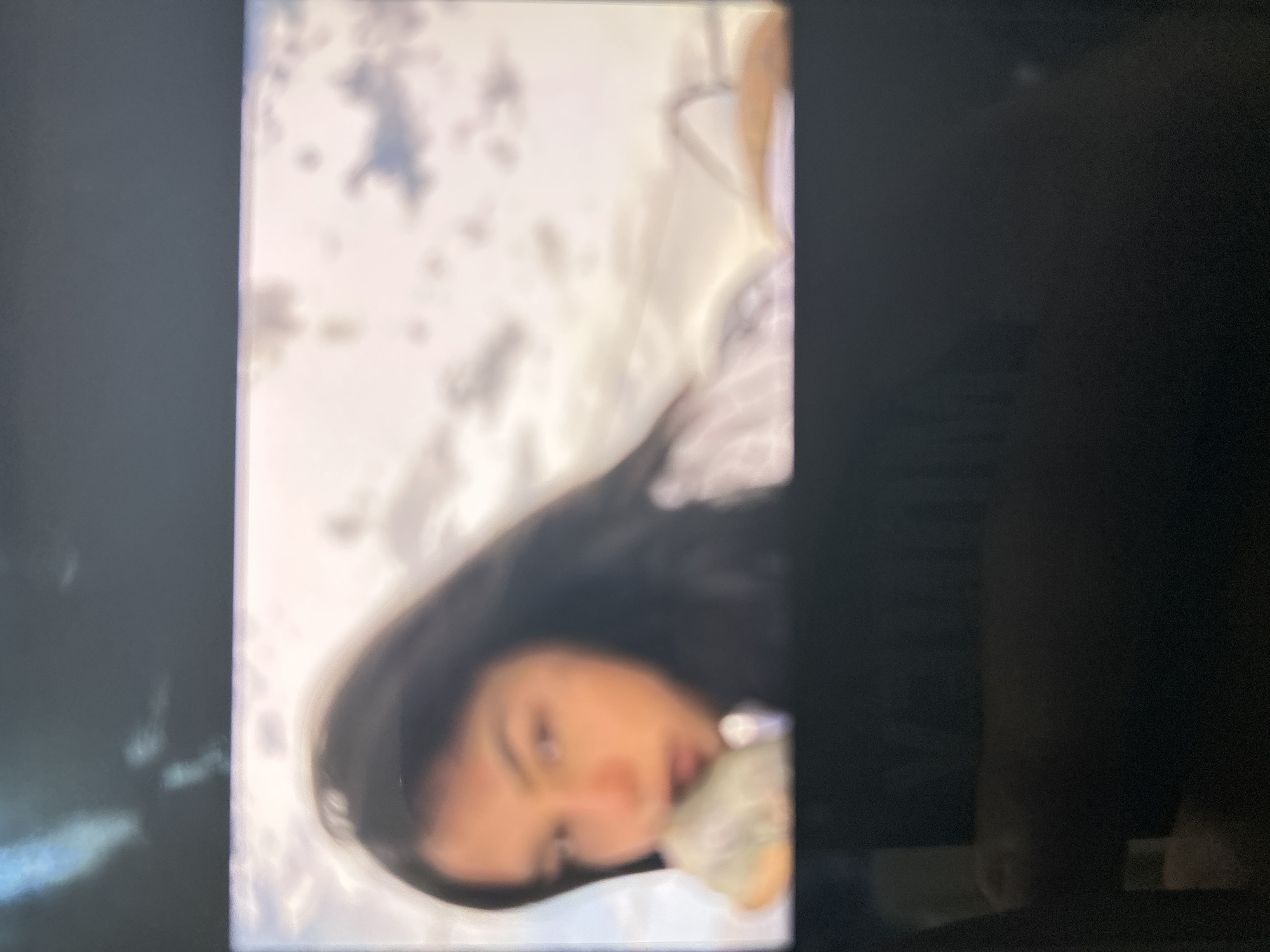 The image is a photograph of a digital display screen, depicting a highly blurred visual content. The top and bottom sections of the overall image show dark, featureless areas consistent with the bezel or frame of a screen, while the central portion displays the primary subject.
The displayed content features a person, likely female, positioned from the chest up against an outdoor background of a cloudy sky. The individual has long, dark hair. Facial features are severely indistinct due to significant blur, but the head is turned slightly to the viewer's left, with a direct or near-direct gaze. An indeterminate, light-colored object is held near the person's mouth or chin, partially obscured by blur, with a hand visible supporting it. The individual appears to be wearing a light-colored, possibly striped, garment. The background consists of a bright, diffuse sky with blurred cloud formations, suggesting an expansive outdoor environment. The scene is situated in the city of Blagoevgrad, Bulgaria.
