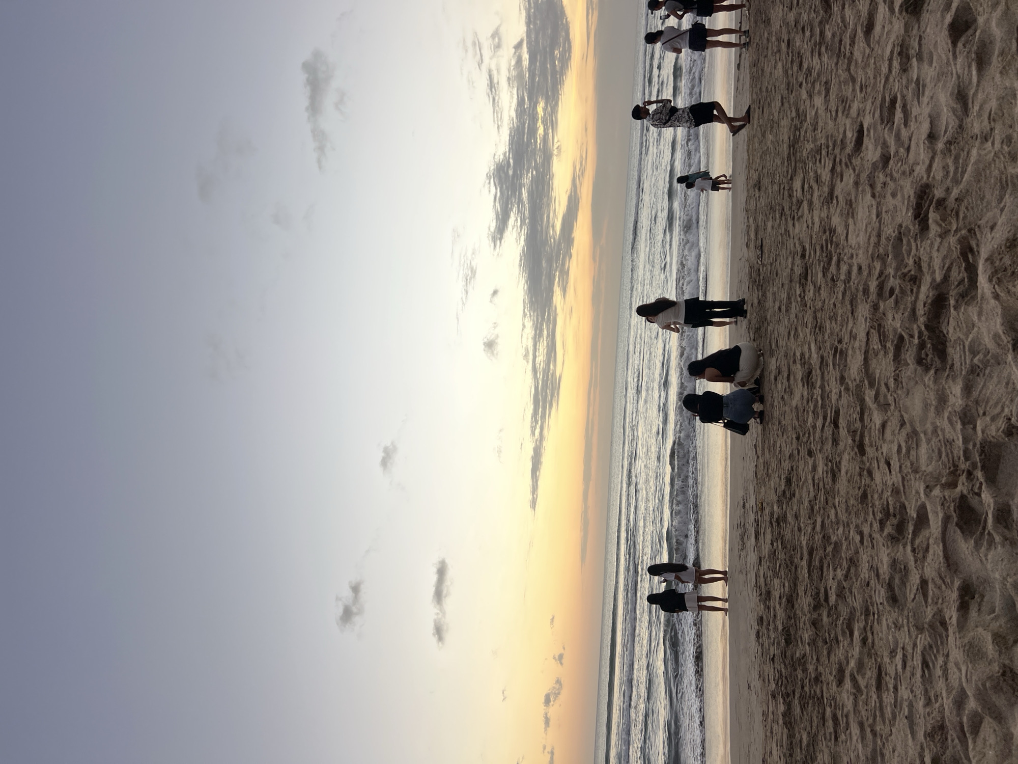 The image depicts a sunset scene at Kuta Beach in Kuta, Indonesia. A group of approximately ten individuals are visible on the sandy shore, with some standing by the water's edge and others further back on the beach. The ocean exhibits moderate waves, and the sky displays the warm hues of a sunset with scattered clouds. The composition is dominated by the sand in the foreground and the sea and sky in the background. The individuals are casually dressed and appear to be enjoying the evening.