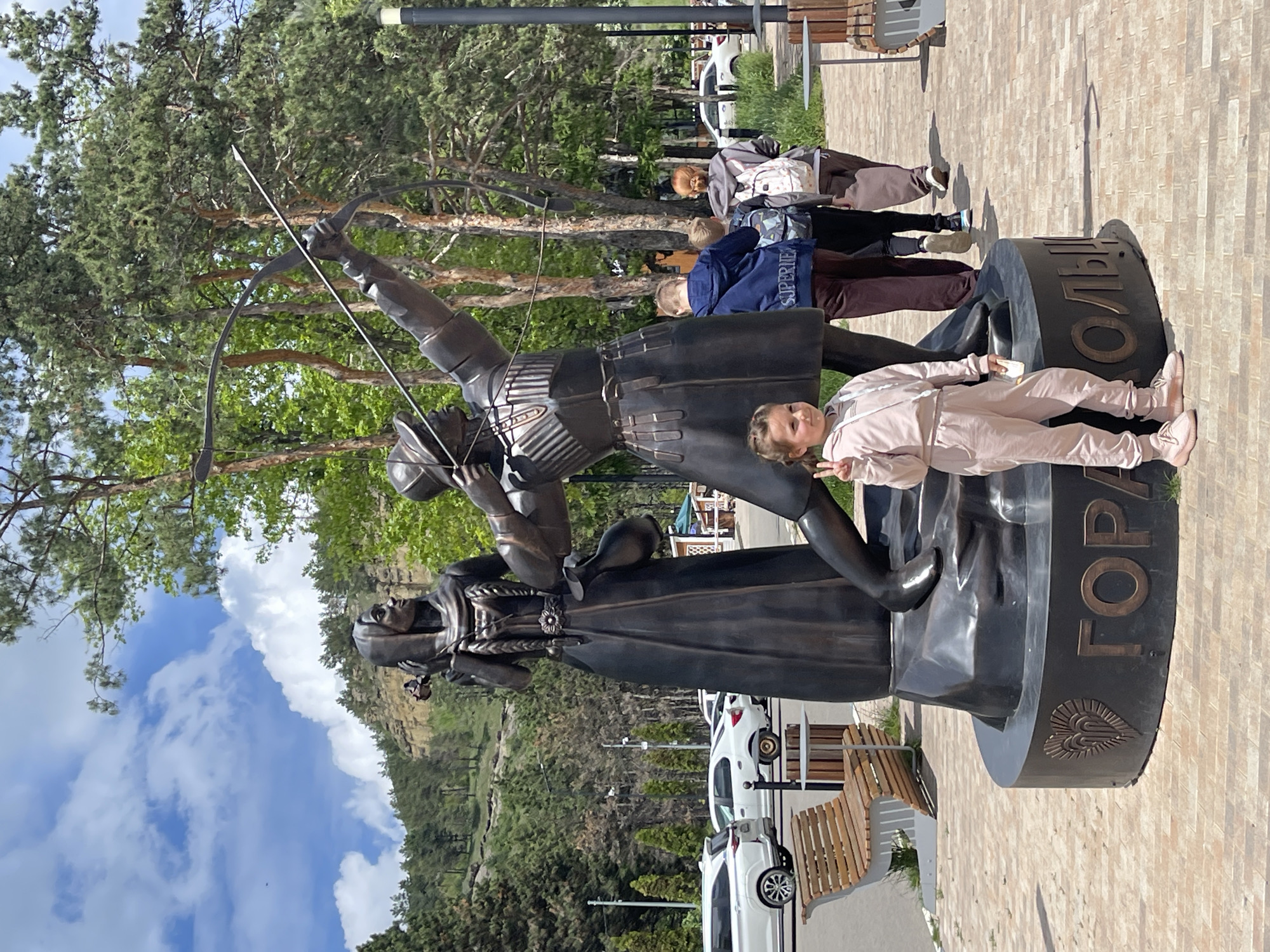The image captures a group of people around a bronze sculpture in Kislovodsk, Russia. The sculpture depicts a male archer aiming an arrow and a woman holding a flower. A girl in a pink jumpsuit is lying on the base of the statue, facing the camera and making a peace sign. Behind the statue, there are several individuals walking, while cars are parked further back. The backdrop includes trees, a cloudy sky, and a view of a hillside. The base of the statue is inscribed with Cyrillic letters. The ground is paved with light brown bricks.