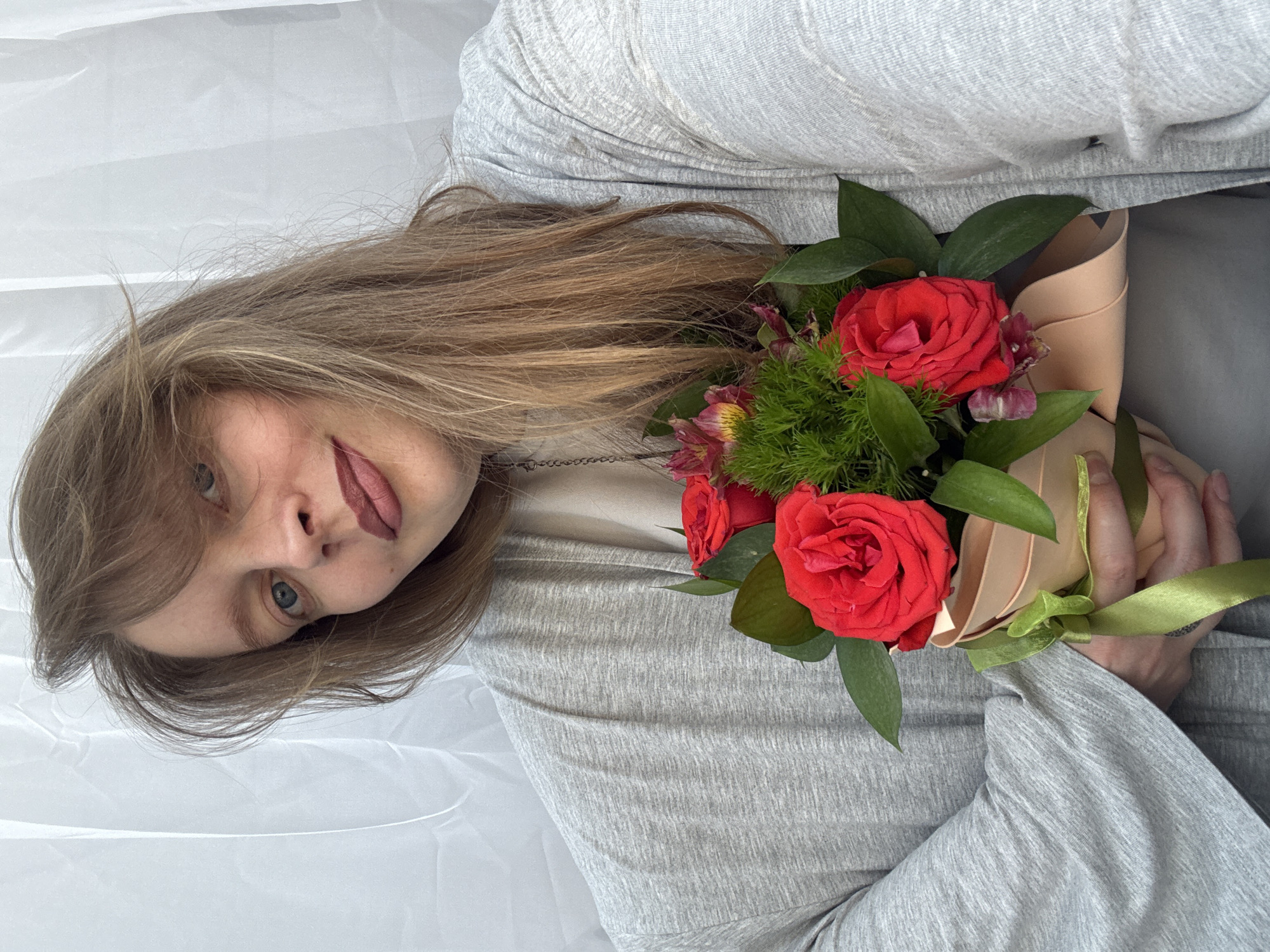 A woman with long, light-brown hair and blue eyes is holding a bouquet of red roses and green foliage. She is wearing a gray long-sleeved shirt or cardigan. The background consists of a white, slightly wrinkled or draped fabric. The image gives no indication of location, especially not Izhevsk, Russia.