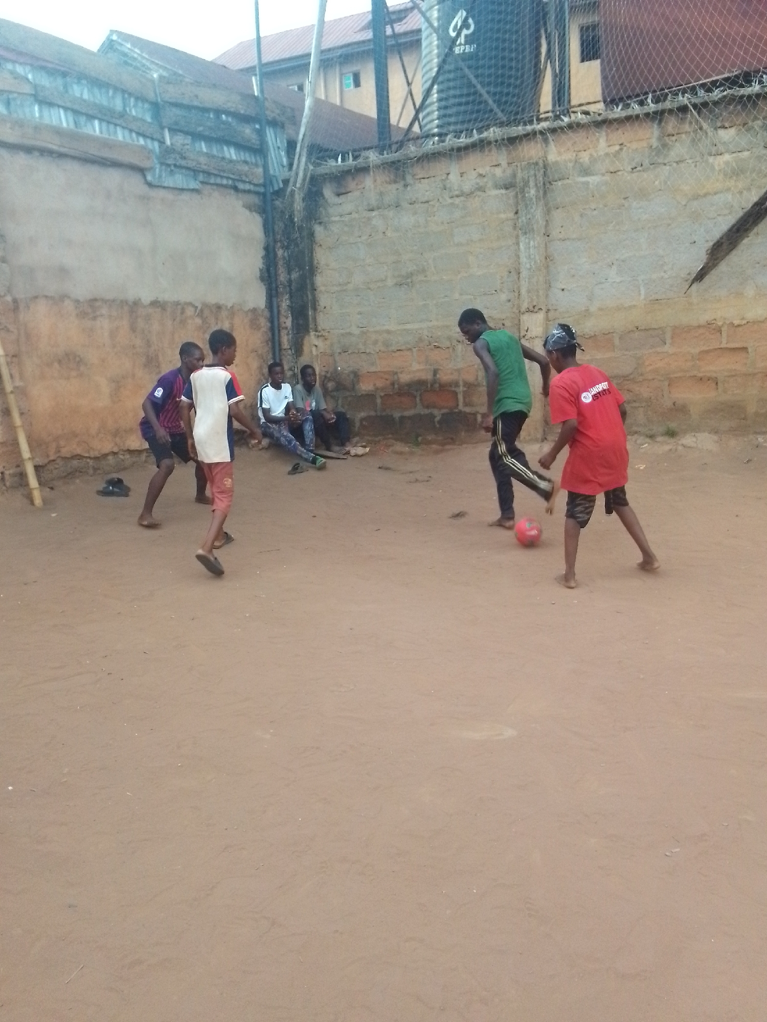 A group of people are playing soccer on a dirt lot in Unknown, Unknown. There are four people watching the game on the side. A water tower and a wall are behind the people.