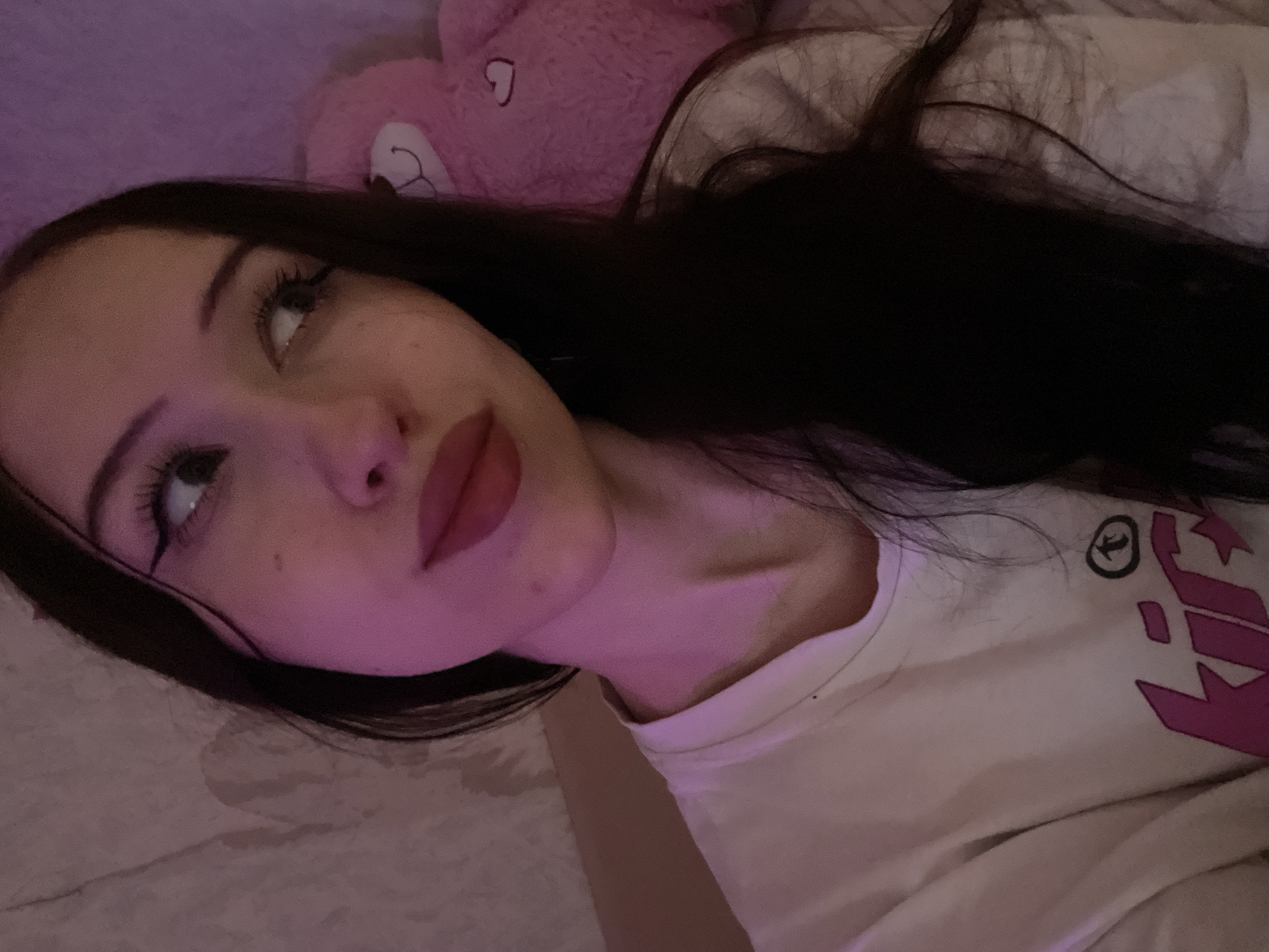 A young woman with long, dark hair and visible makeup is captured in a close-up, over-the-shoulder selfie. She is wearing a white t-shirt with pink lettering and graphics. To her right, a large, pink stuffed teddy bear is partially visible. The lighting in the scene is predominantly pink. The background appears to be a textured wall or fabric, possibly in a bedroom or living space. There is no discernible location within Votkinsk, Russia, or any other specific city.