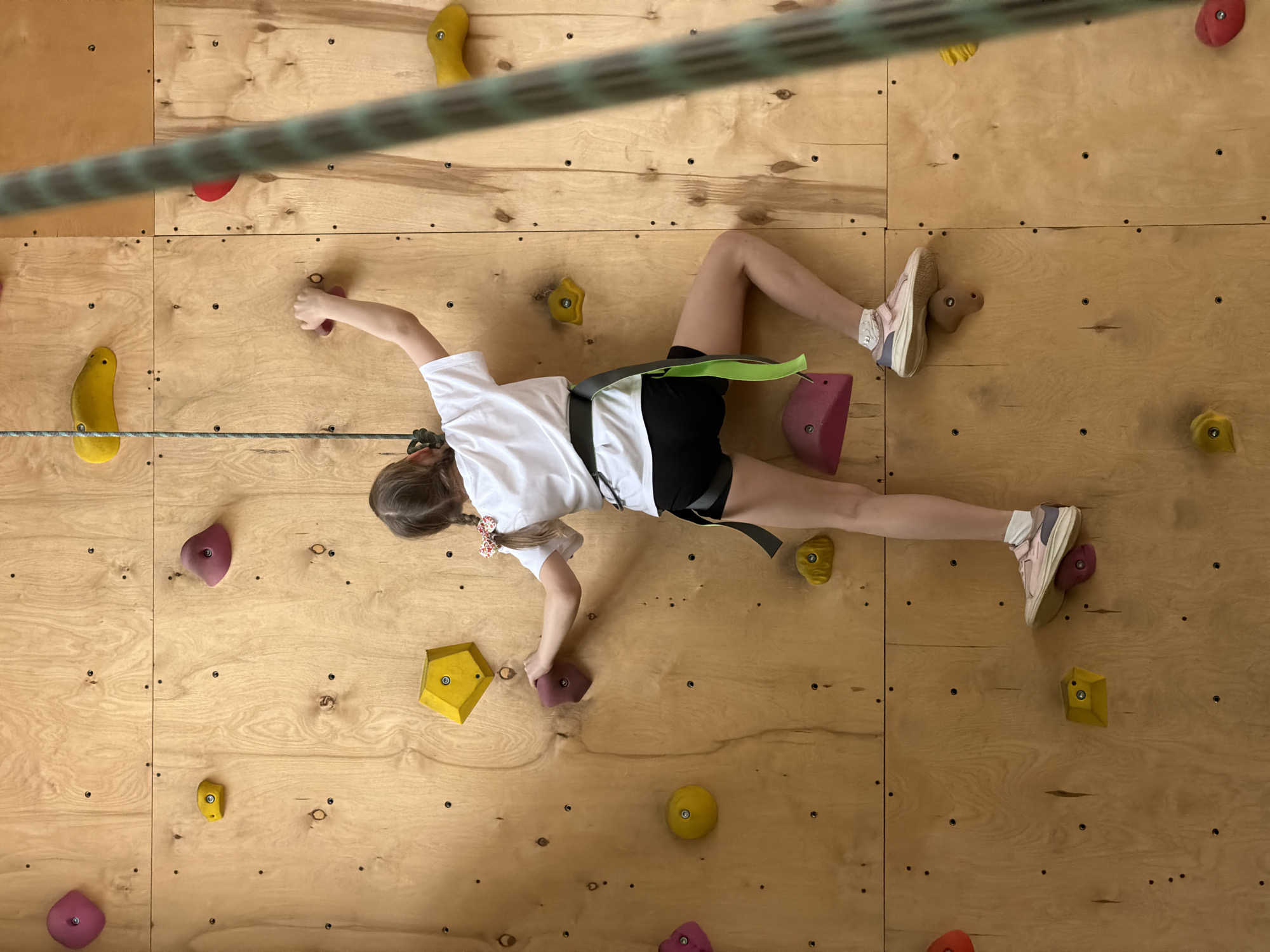 A girl is scaling a climbing wall. She is wearing a white T-shirt, black shorts, pink and white sneakers, and a climbing harness. The climbing wall is made of plywood and has various colored holds attached to it. A rope is visible across the top of the image. The location is Izhevsk, Russia.