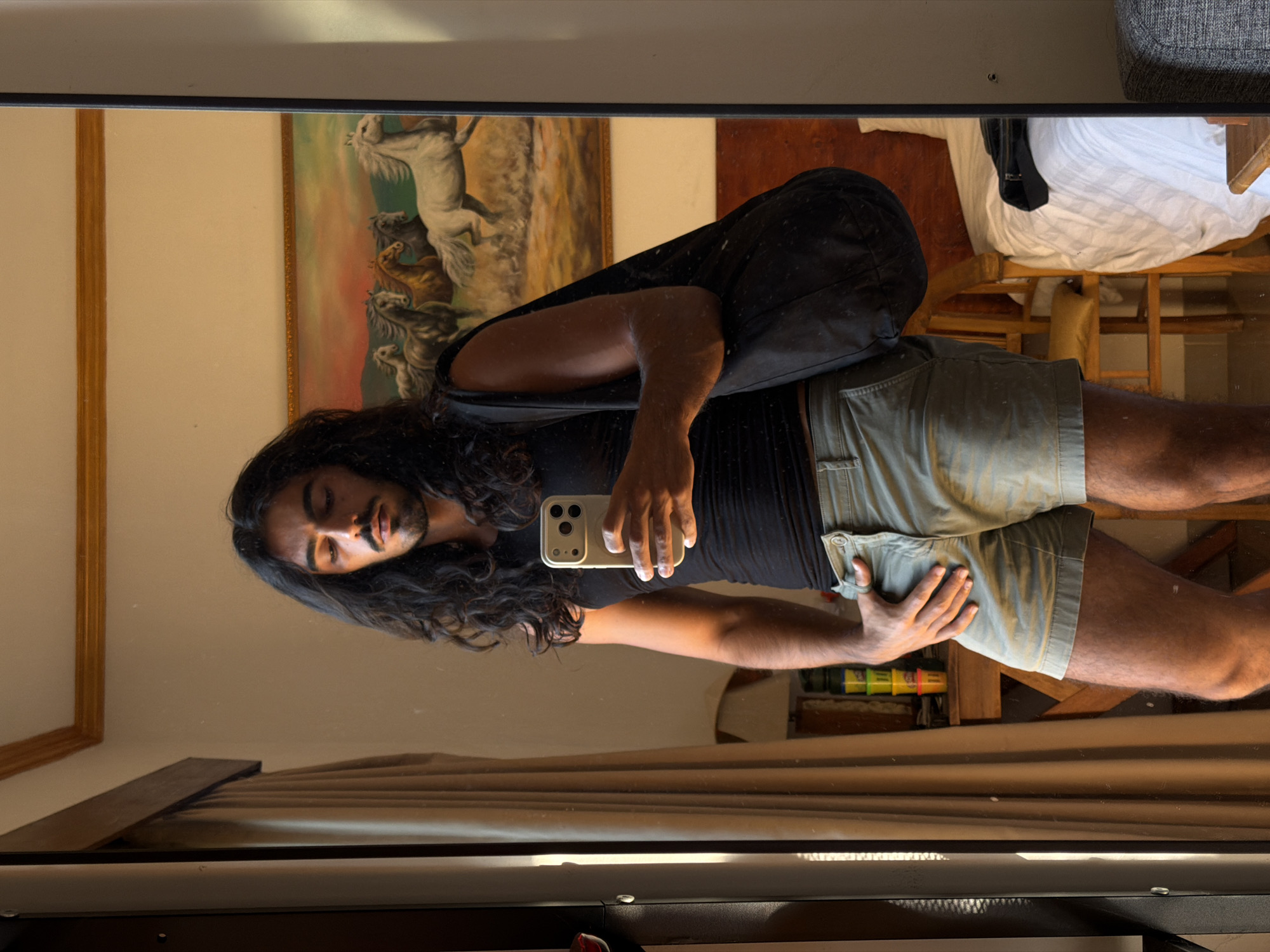 A person is visible in a mirror selfie, positioned against a wall where a framed painting is displayed. The painting depicts white horses in a landscape. The person is wearing a black sleeveless shirt and light green shorts, with a black bag slung over their shoulder. Their hair is long and dark. They are holding a cell phone, presumably to take the picture. A partially visible bed with white sheets is in the background to the right. The location is North Kuta, Indonesia.