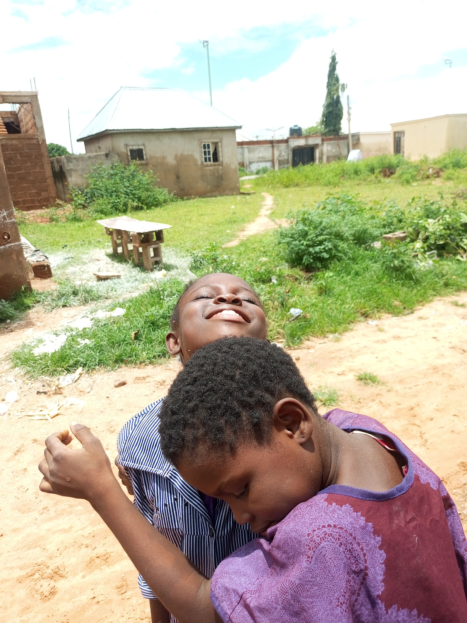 In Tempure, Nigeria, two young children are embracing. The child in the foreground, wearing a purple garment with a patterned design, rests their head on the shoulder of the child behind them, who is wearing a blue and white striped shirt. The child with their head down appears to be asleep or resting, with their eyes closed and their face turned away from the camera. The child behind them has their eyes closed and their head tilted back, with a joyful expression. The background of the image shows a residential area with simple, unpainted buildings, some under construction, and a tall, slender tree. The ground is a mix of sand and grass, with scattered debris. The sky is bright blue with scattered clouds, indicating a sunny day. A small, simple wooden table with a metal top is visible in the middle ground. The overall scene suggests a casual outdoor moment in a rural or semi-rural Nigerian setting.