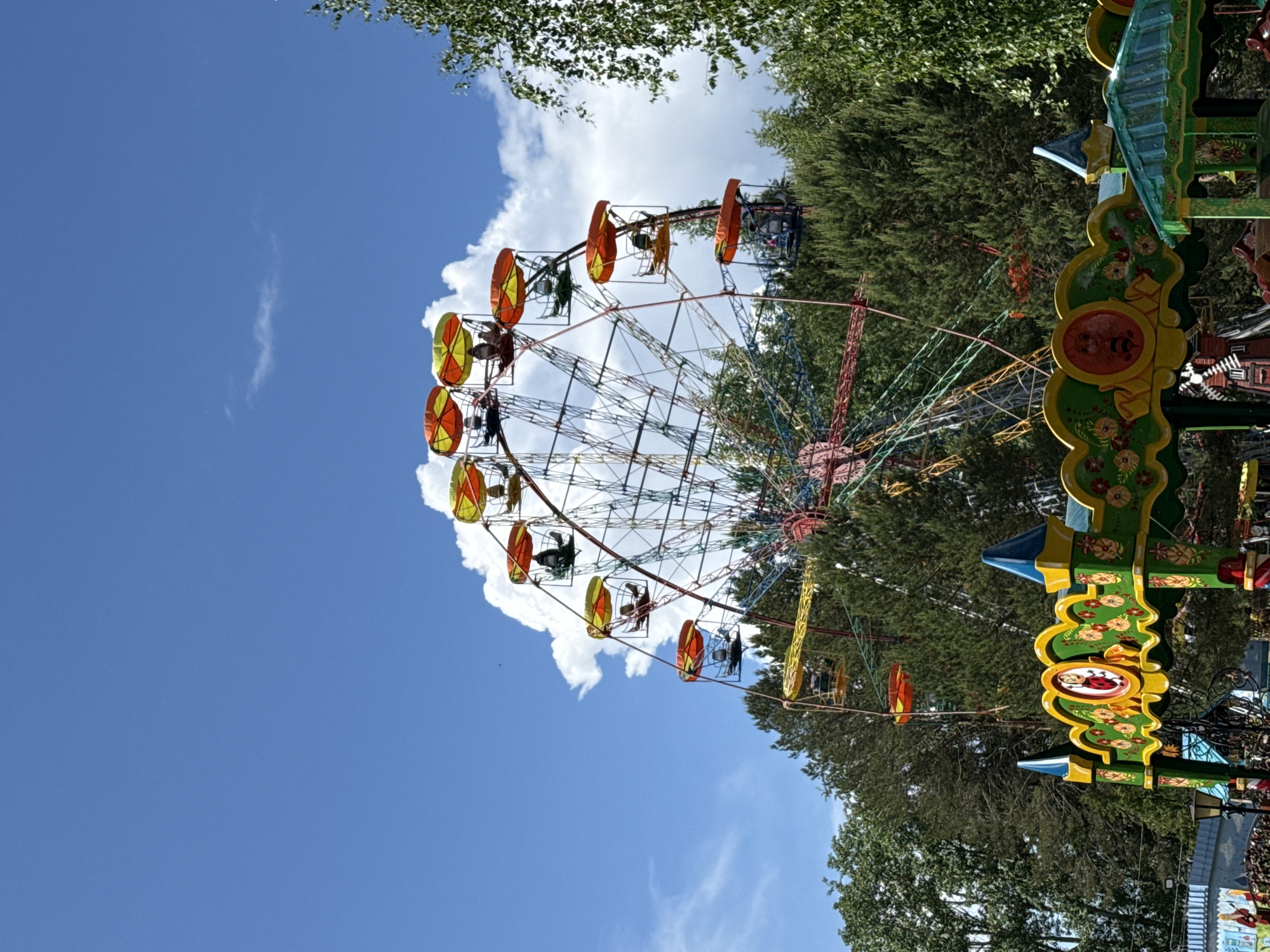 The image shows a colorful ferris wheel in Chaikovsky, Russia. The ferris wheel is partially obscured by trees and a large white cloud. The sky is blue with a few scattered clouds. The ferris wheel has open carriages with yellow and orange roofs. A decorative, painted structure is visible on the right side of the frame, also partially obscured by trees.