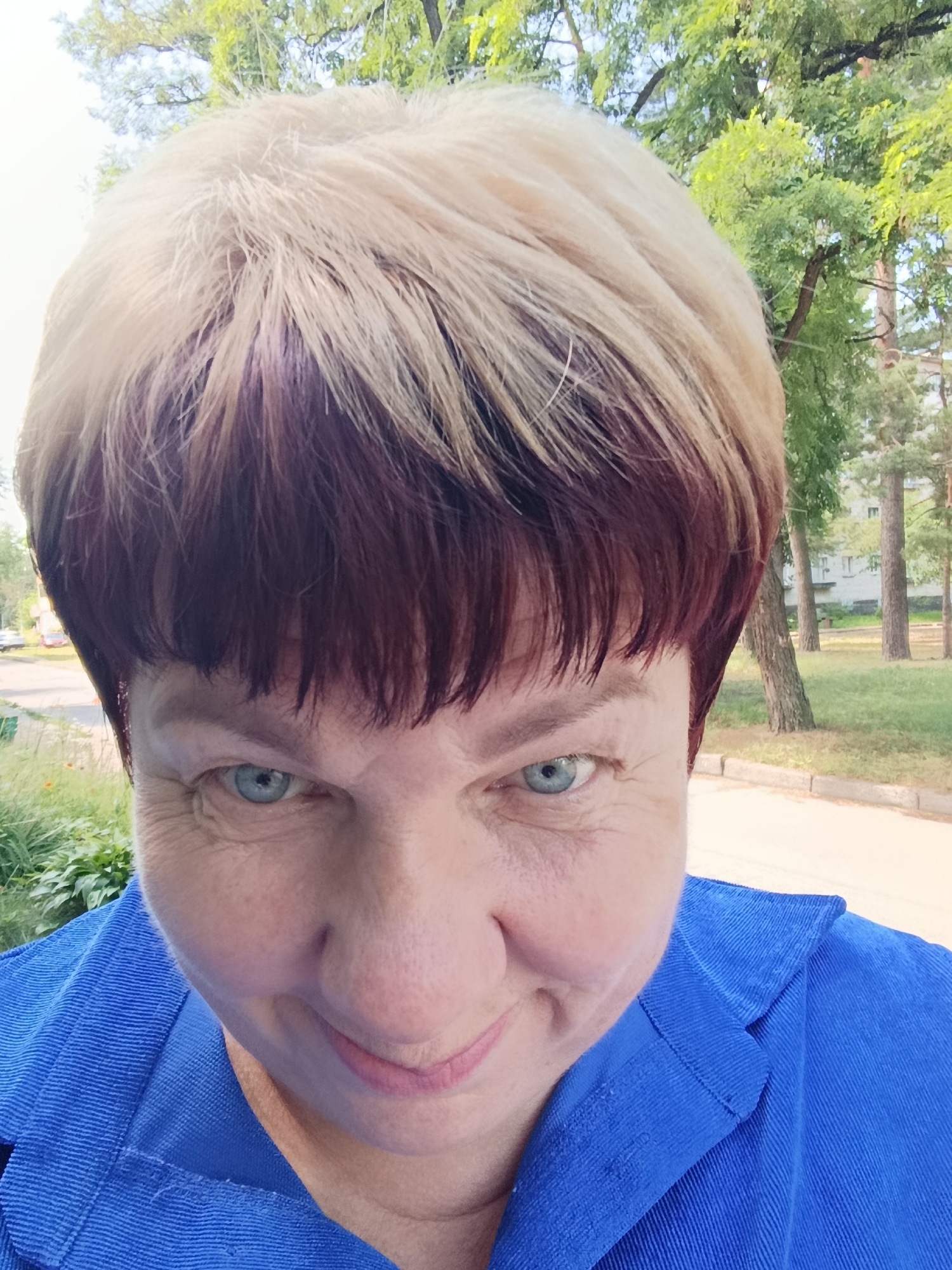 A close-up, waist-up selfie features a woman with short, two-toned hair, blonde on top and dark reddish-brown underneath. Her head is tilted slightly downwards, and she is looking directly at the camera with light blue eyes. She is wearing a vibrant blue jacket with visible stitching and corduroy texture. The background appears to be an outdoor urban park setting with lush green trees, grass, and a paved walkway. In the distance, a few buildings and parked cars are visible, suggesting the location is Svietlahorsk, Belarus.