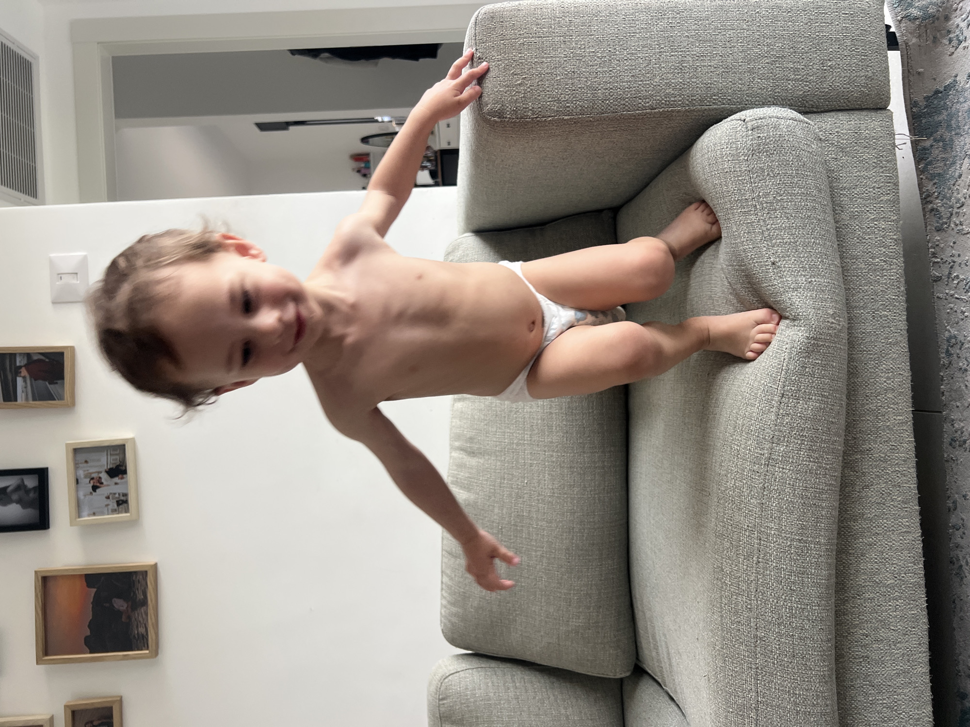 A small child, wearing a diaper, is positioned atop a gray upholstered sofa. The child is reaching toward the back of the sofa with one hand and has their feet on the armrest. In the background is a white wall decorated with several framed pictures. A doorway is visible, leading to another room. The location is likely a residence, but Kiryat Bialik, Israel, cannot be confirmed based on the visual information provided.