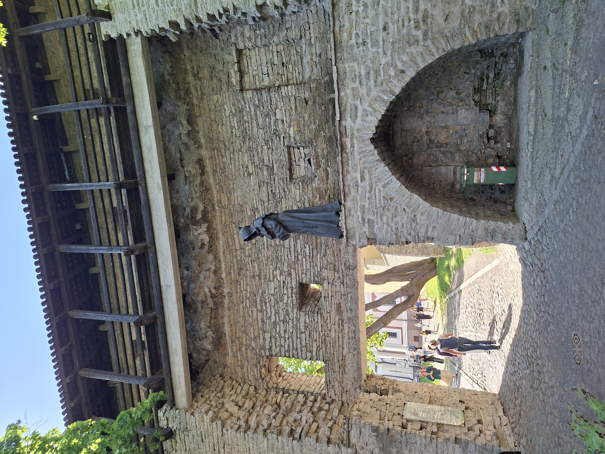A sunlit outdoor scene in Tallinn, Estonia, depicts a historic stone archway and wall structure, likely part of the city's fortifications. The main focal point is a dark, cloaked figure or statue affixed to the rugged stone wall, positioned above the arched passageway. Above this wall, a wooden open-beam structure is visible, suggesting a walkway or part of a defensive gallery.
Through the stone archway, a cobblestone street stretches into the distance under bright daylight. Several people are visible on the street: one person in dark clothing walks away from the archway in the immediate foreground, while others are further down, some seated or standing near buildings. A large tree trunk and green foliage are also seen through the archway, casting dappled shadows.
Inside a niche within the archway, a green trash bin with red and white striped markings is placed. Text on the bin includes "VÄIKEPRÜGI", "SÄTI KORDA", and "TALLINN". A plaque with unreadable text is also visible on the lower section of the stone wall to the right of the archway. The sky is clear blue, indicative of good weather, and strong shadows suggest the time of day is around midday or early afternoon. The overall impression is a bustling yet historic street scene.