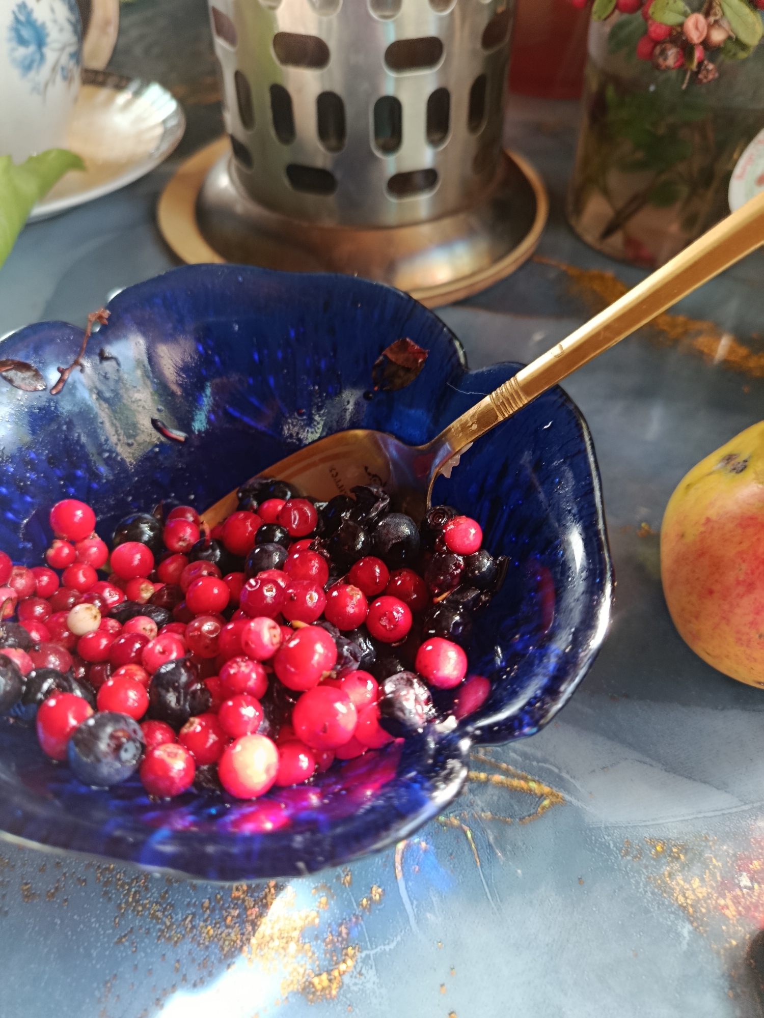 The image shows a close-up of a blue ceramic bowl filled with red berries and blueberries. A gold spoon rests in the bowl, partially submerged in the berries and juice. To the right of the bowl, a partially visible yellow and red fruit, likely an apple or pear, rests on a blue surface. In the background, a stainless steel object, possibly a tea infuser, stands on a wooden coaster. A glass jar with red berries and green leaves is also visible behind the infuser. A white teacup with blue floral patterns is on a saucer to the left of the infuser. The overall setting appears to be a tabletop with a textured, possibly marble or granite, surface. The image does not provide any specific information about the location in the city of Верея, Russia.