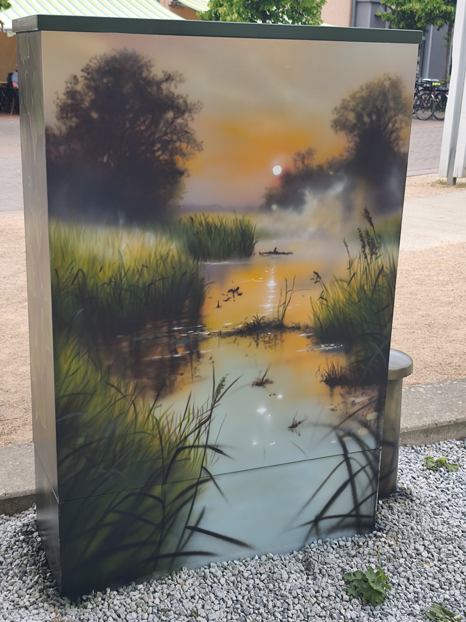 A utility box is located on the street in Unknown, Unknown, and is painted with a landscape scene of a marsh at sunset. Tall grass and reeds are visible in the foreground and midground, with a body of water reflecting the orange and yellow hues of the setting sun. Trees are silhouetted against the sky in the background, adding depth to the landscape. The box is surrounded by small gravel.