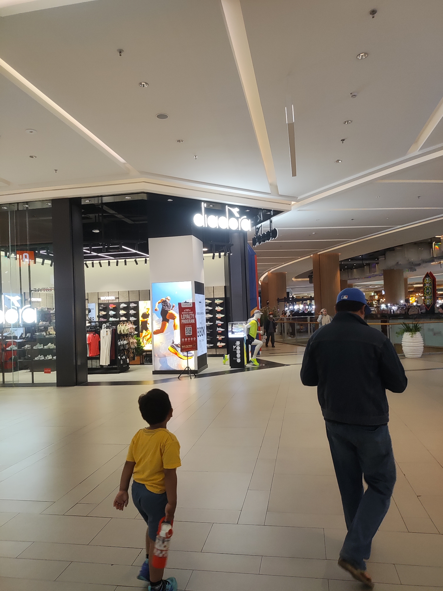 The image captures an indoor scene within a mall, likely located in Lambangjaya, Indonesia. In the foreground, a young boy wearing a yellow t-shirt and blue shorts walks away from the camera, holding a red and white water bottle. To the right and further back, a man in a dark jacket and blue jeans walks away from the camera, wearing a blue baseball cap.
The environment appears to be a spacious shopping mall with a high ceiling featuring modern lighting fixtures and recessed lighting. In the midground, a Diadora store is visible, showcasing athletic footwear and apparel on display. A large digital advertisement for a loyalty program is prominently placed outside the store. Other shoppers can be seen in the background, as well as decorative plants and mall architecture. The floor is tiled with a light-colored material.