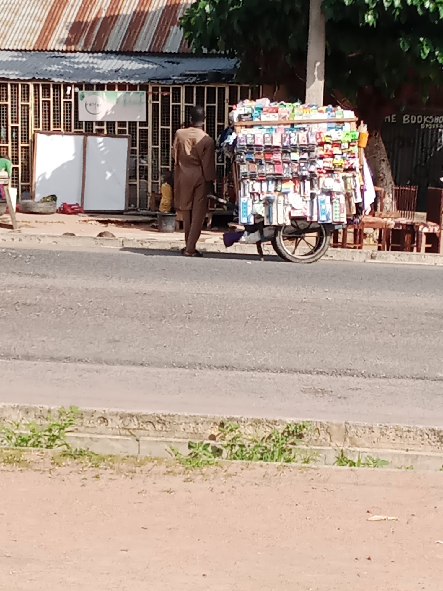 The image captures a street scene in Jalingo, Nigeria, where a vendor stands beside a cart laden with various items, likely for sale. The vendor, dressed in a brown tunic and trousers, appears to be moving the cart, which is a mobile stall featuring an array of packaged goods, possibly including phone accessories and small electronics, displayed on shelves. The cart has a single large wheel, suggesting it's designed for easy maneuverability.
Behind the vendor and cart, a building with a corrugated metal roof and barred windows forms the backdrop. A sign with a logo that appears to be "fair" is visible on the building's facade. To the right, part of a sign reads "NE BOOKSHOP," indicating another establishment nearby. The street in the foreground is paved and shows signs of wear. Vegetation, including grass, grows along the edge of the road, suggesting a semi-urban or transitional area within the city. The overall lighting suggests it is daytime.